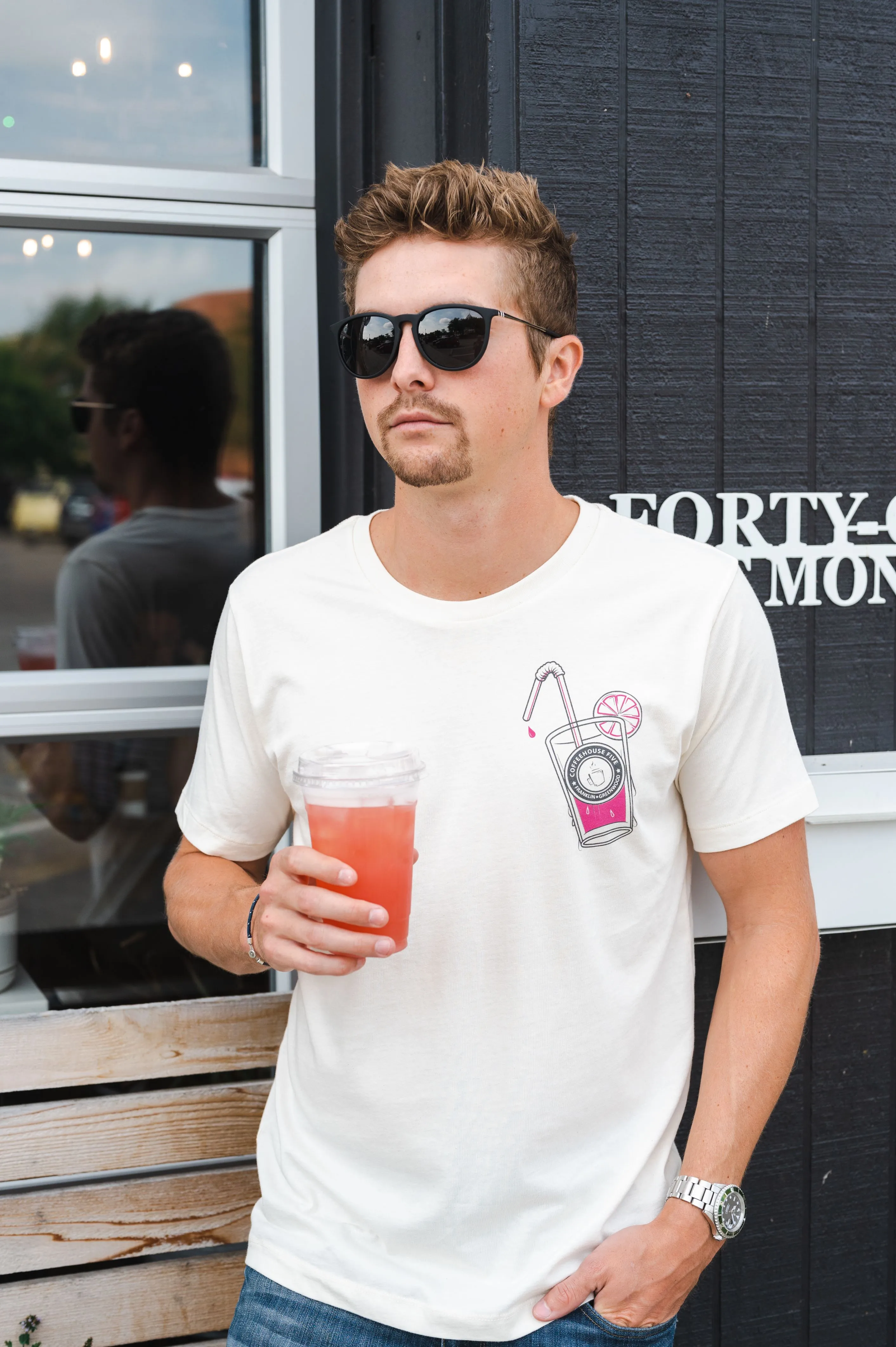 Man in sunglasses and white t-shirt with graphic design holding a pink beverage, standing in front of a dark building with window reflections.