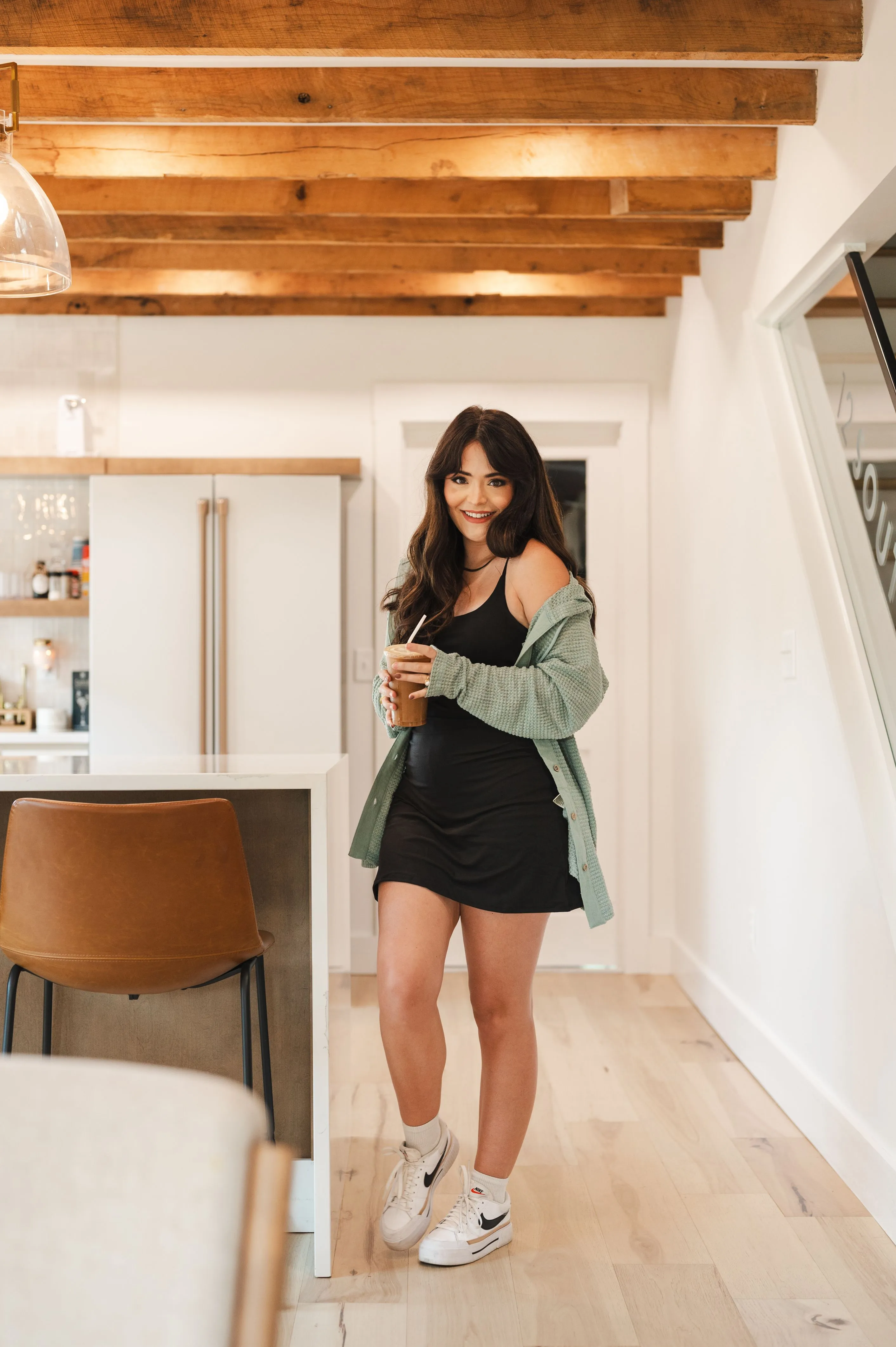 Smiling woman standing in a modern kitchen, holding a cold beverage, wearing a black dress with a green cardigan and white sneakers.
