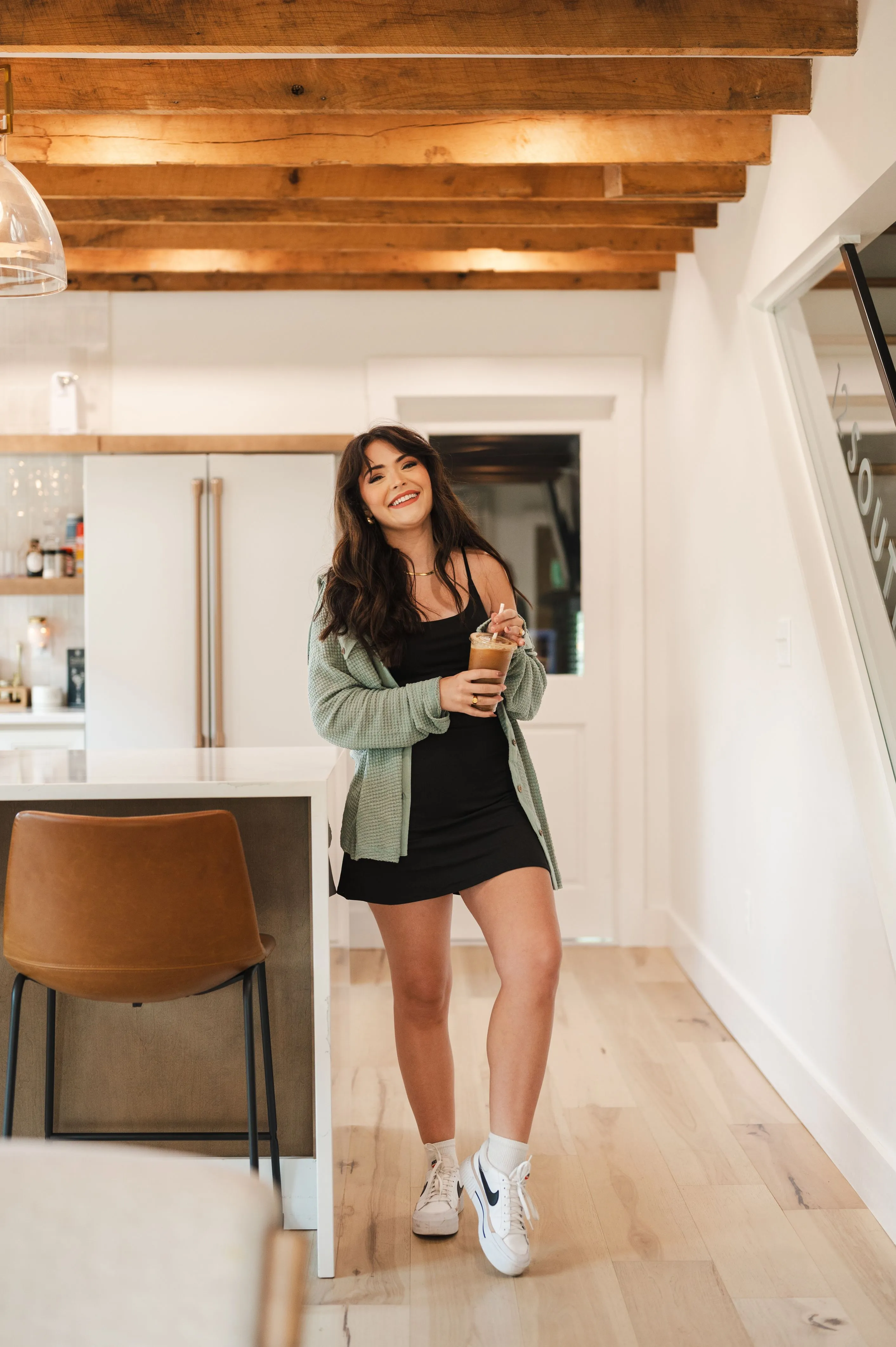 A smiling woman holding a coffee cup standing in a modern kitchen with wooden beams on the ceiling.