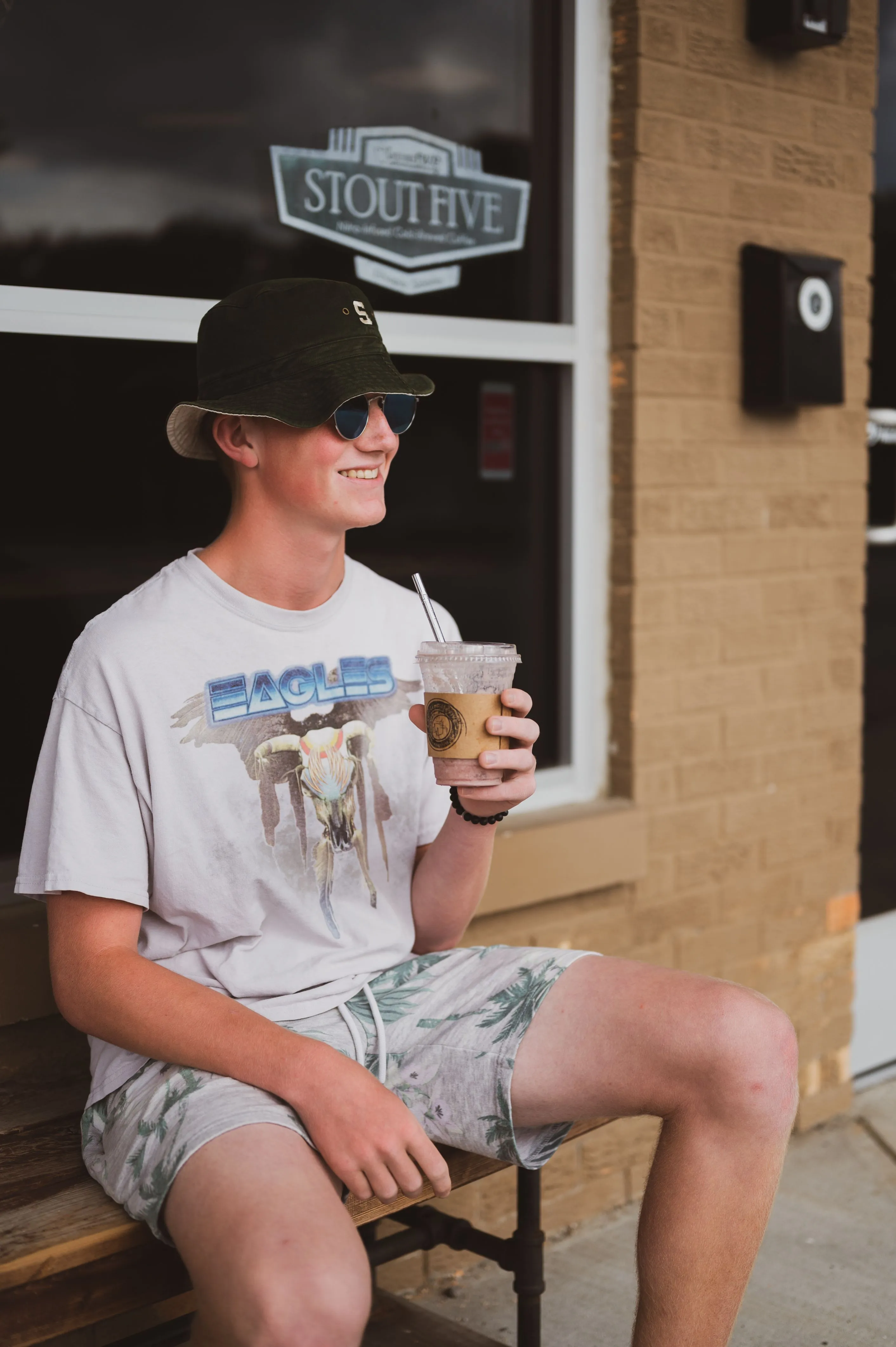 Smiling young person sitting on a bench outside a café, wearing a bucket hat and sunglasses, holding an iced coffee drink.