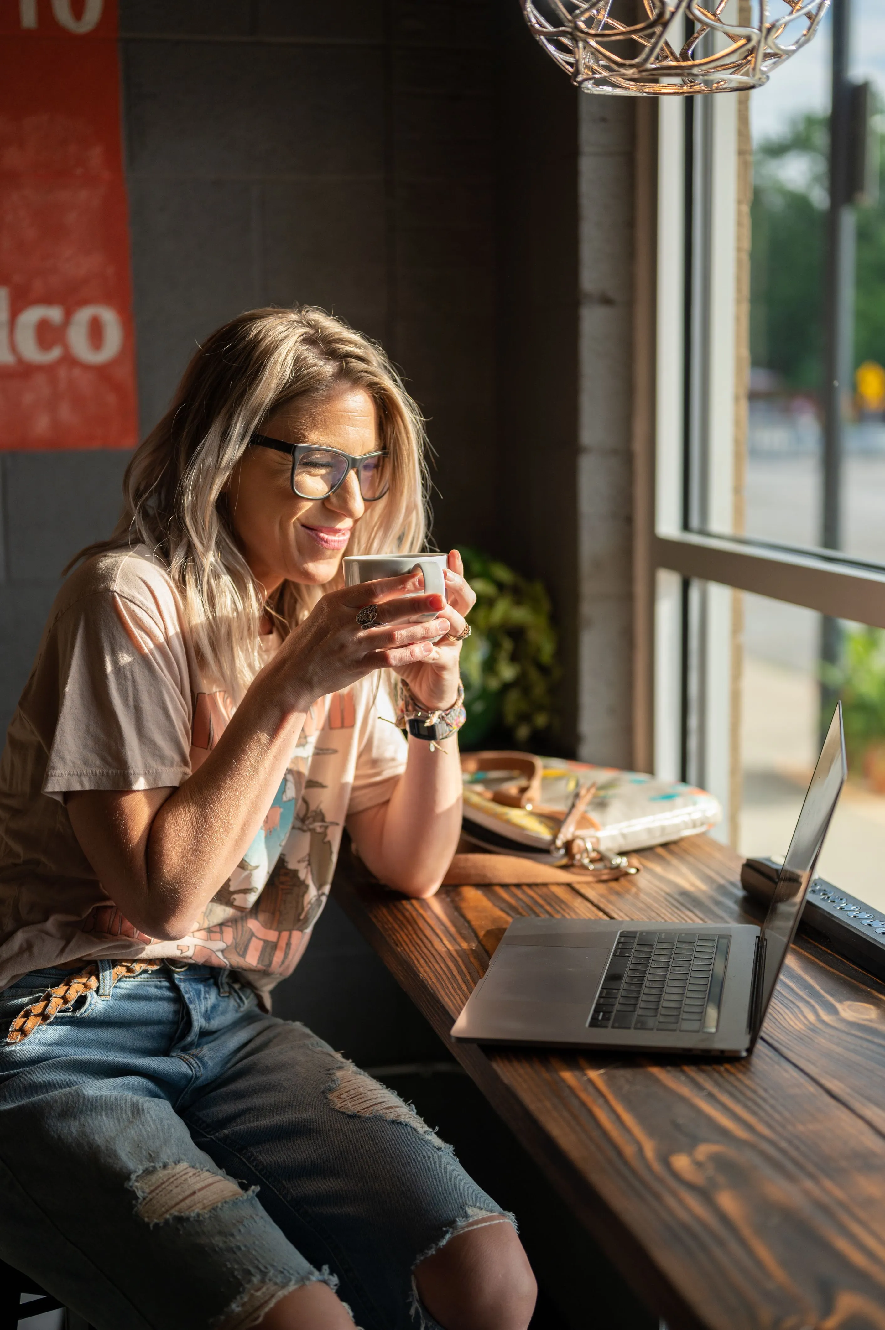 Woman smiling while holding a cup of coffee at a table with a laptop and personal items, in a sunlit cafe.
