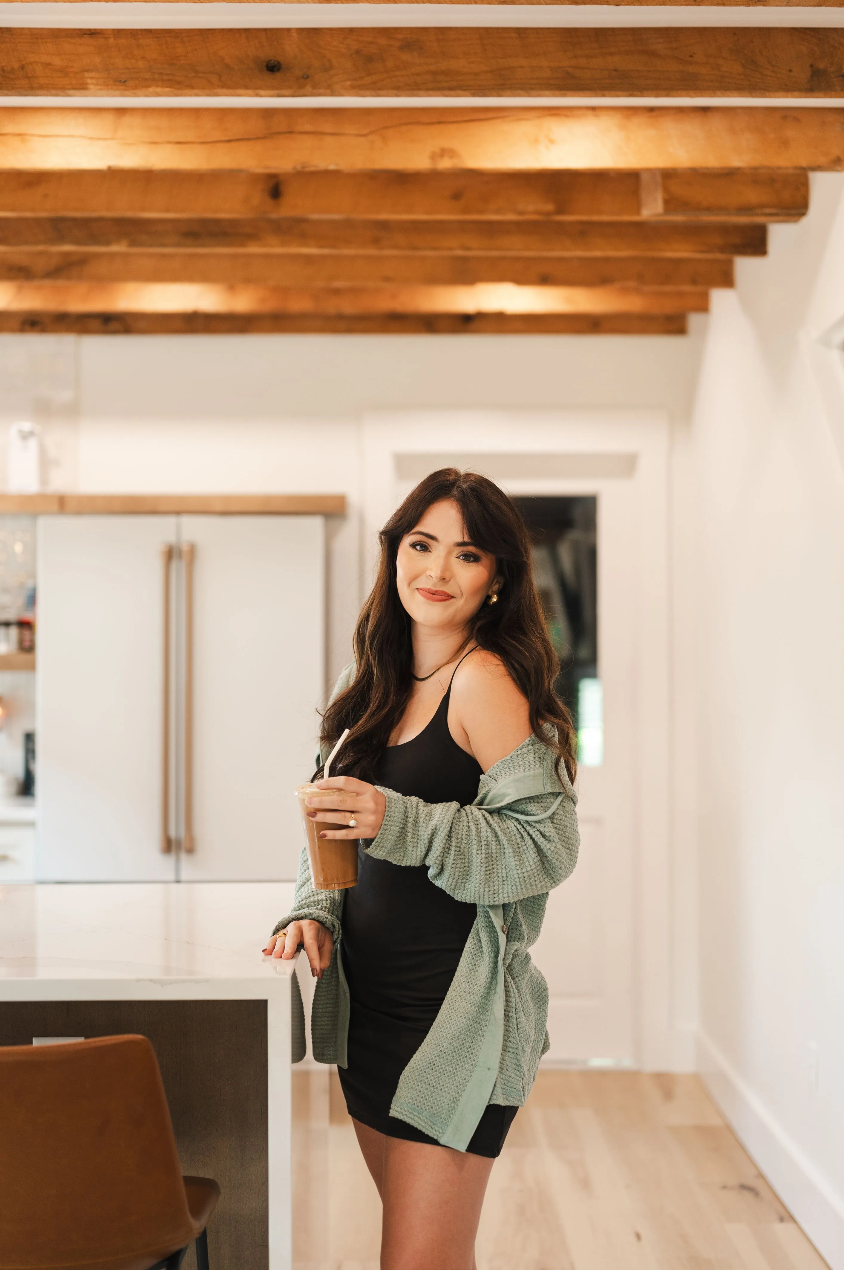 Woman standing in a modern kitchen holding a cold beverage, wearing a black dress and a green cardigan, with exposed ceiling beams above.