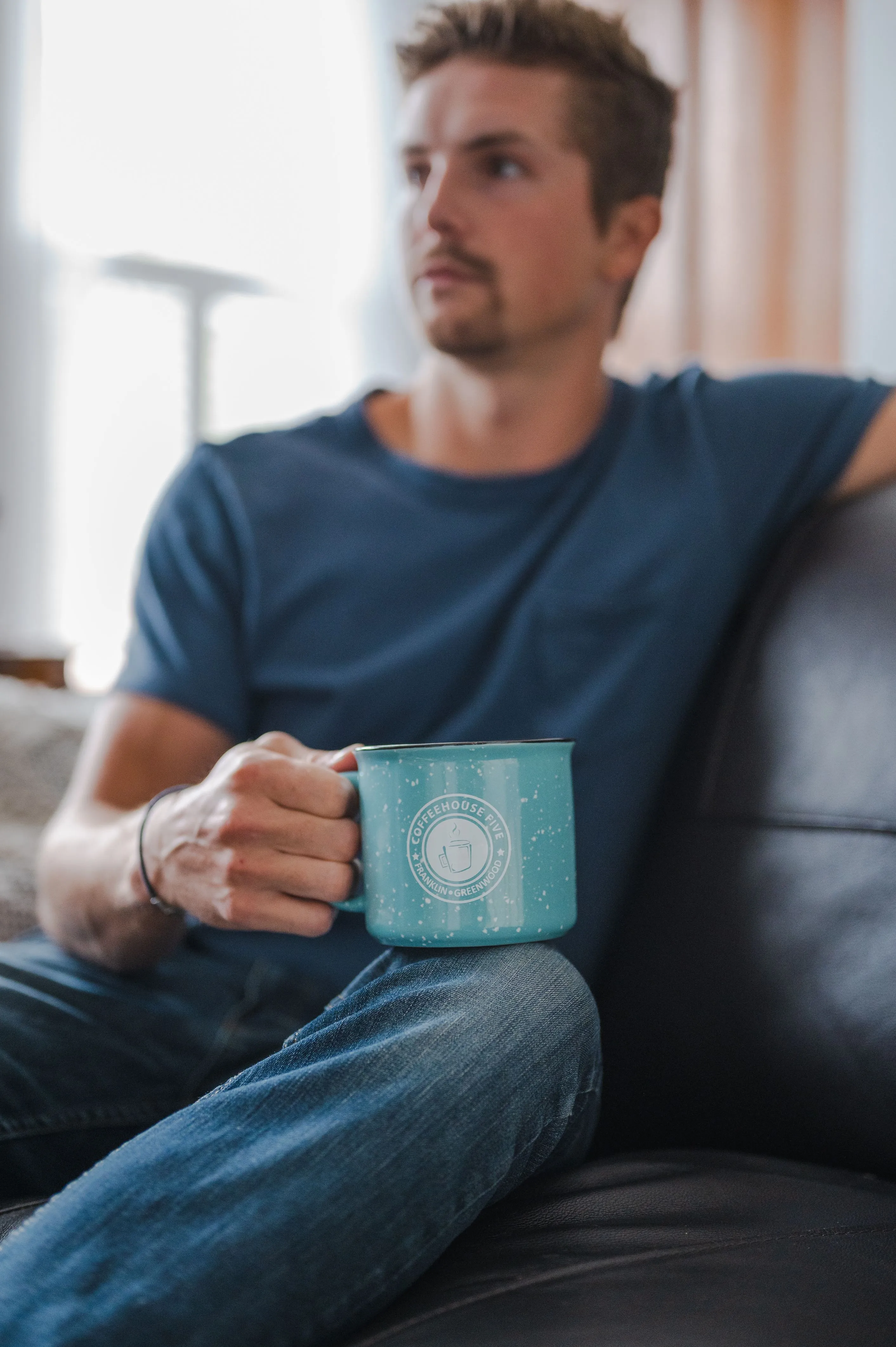 Man sitting on a couch holding a blue mug, looking thoughtfully off to the side.