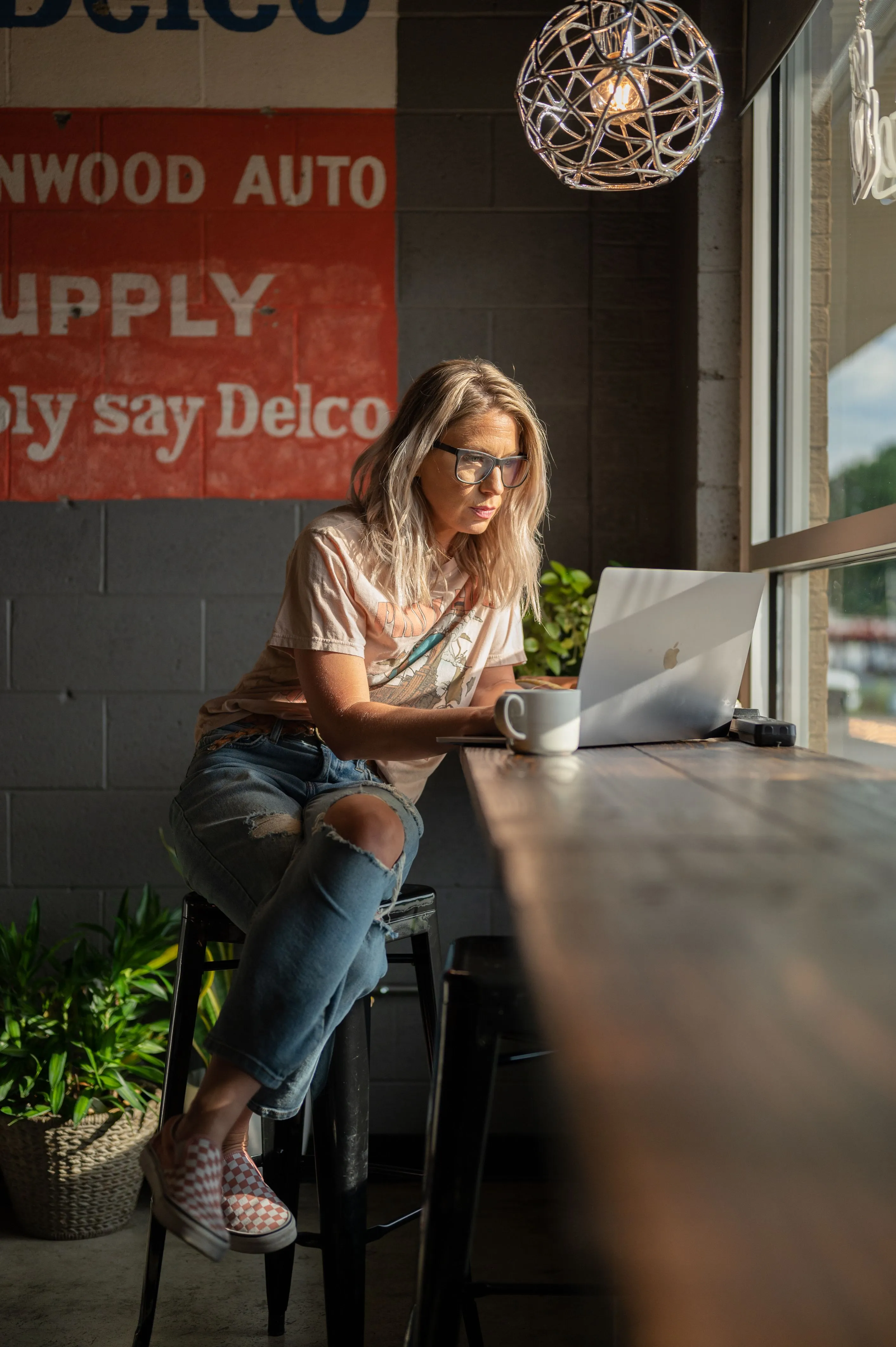 Woman sitting at a cafe table working on a laptop with a coffee cup, an industrial-style pendant light above and a vintage auto supply sign in the background.