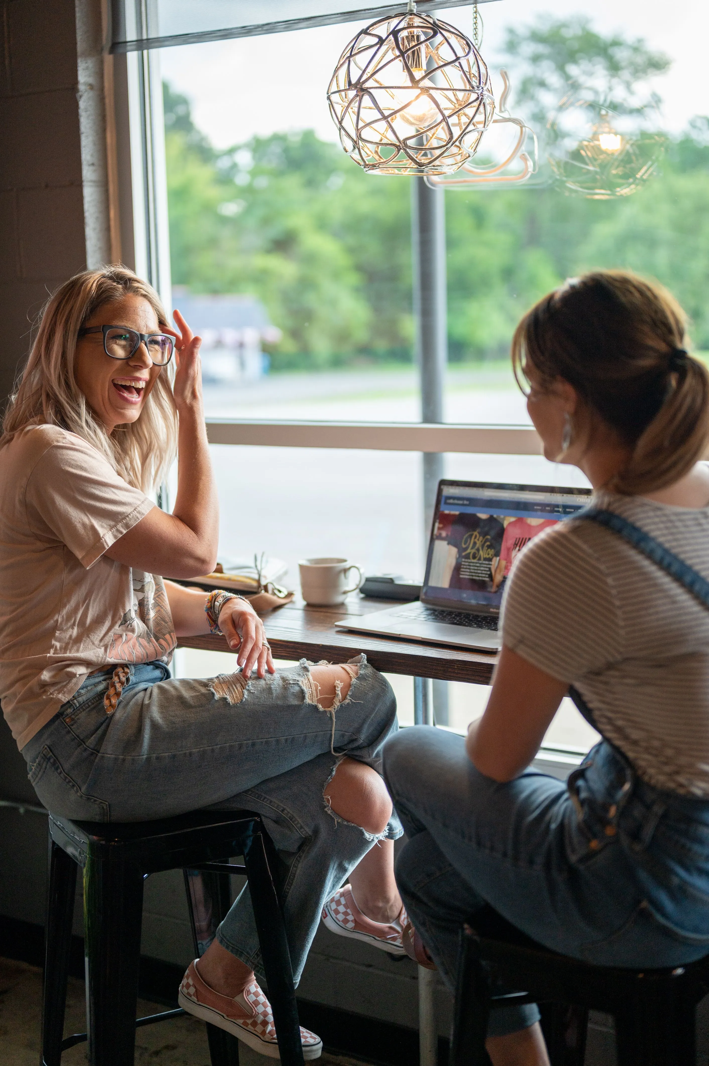 Two women seated at a cafe table beside a window, with one laughing and gesturing while the other looks at a laptop on the table.