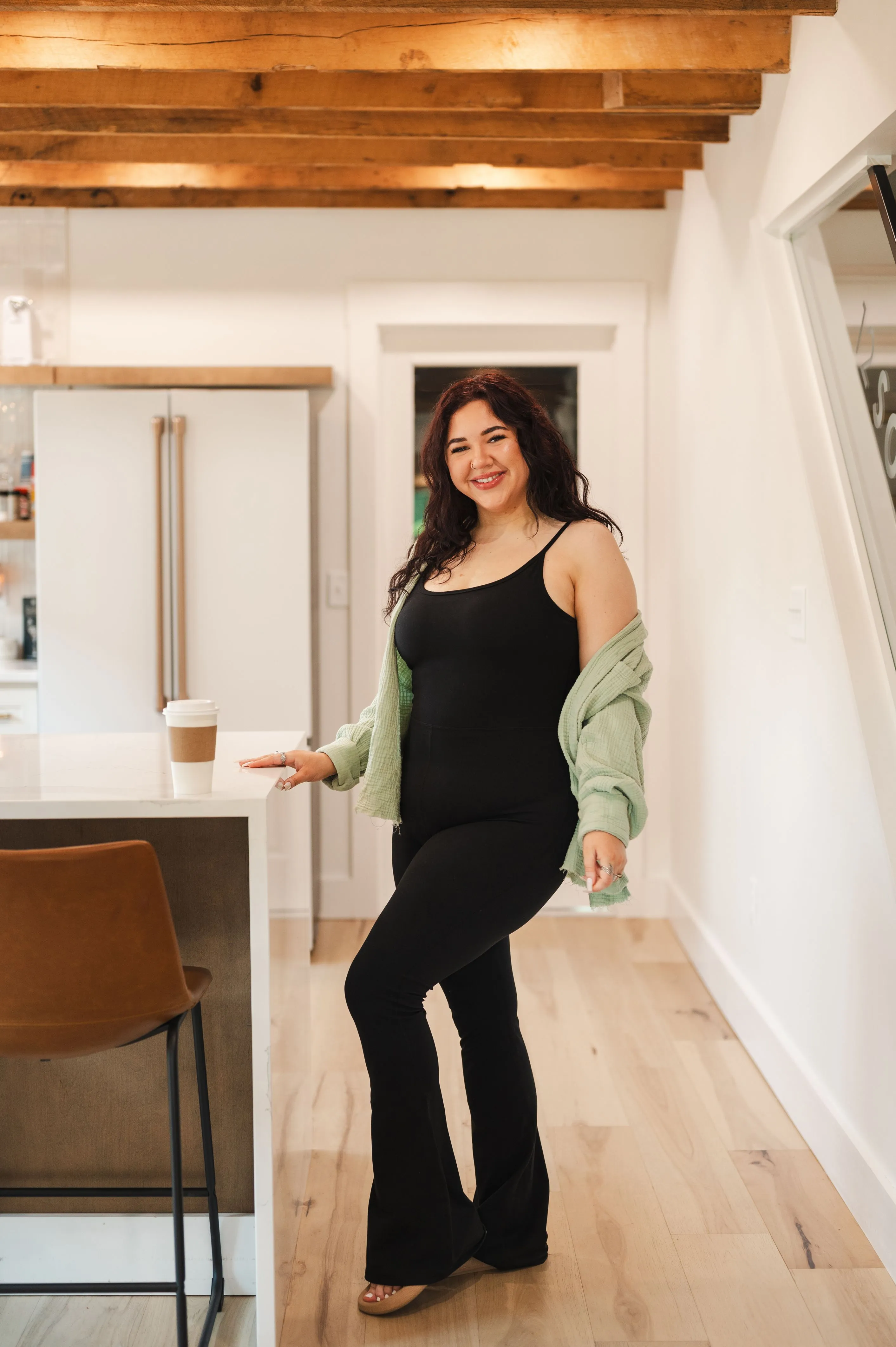 A smiling woman standing in a kitchen, wearing a black outfit with a green cardigan, next to a counter with a coffee cup.