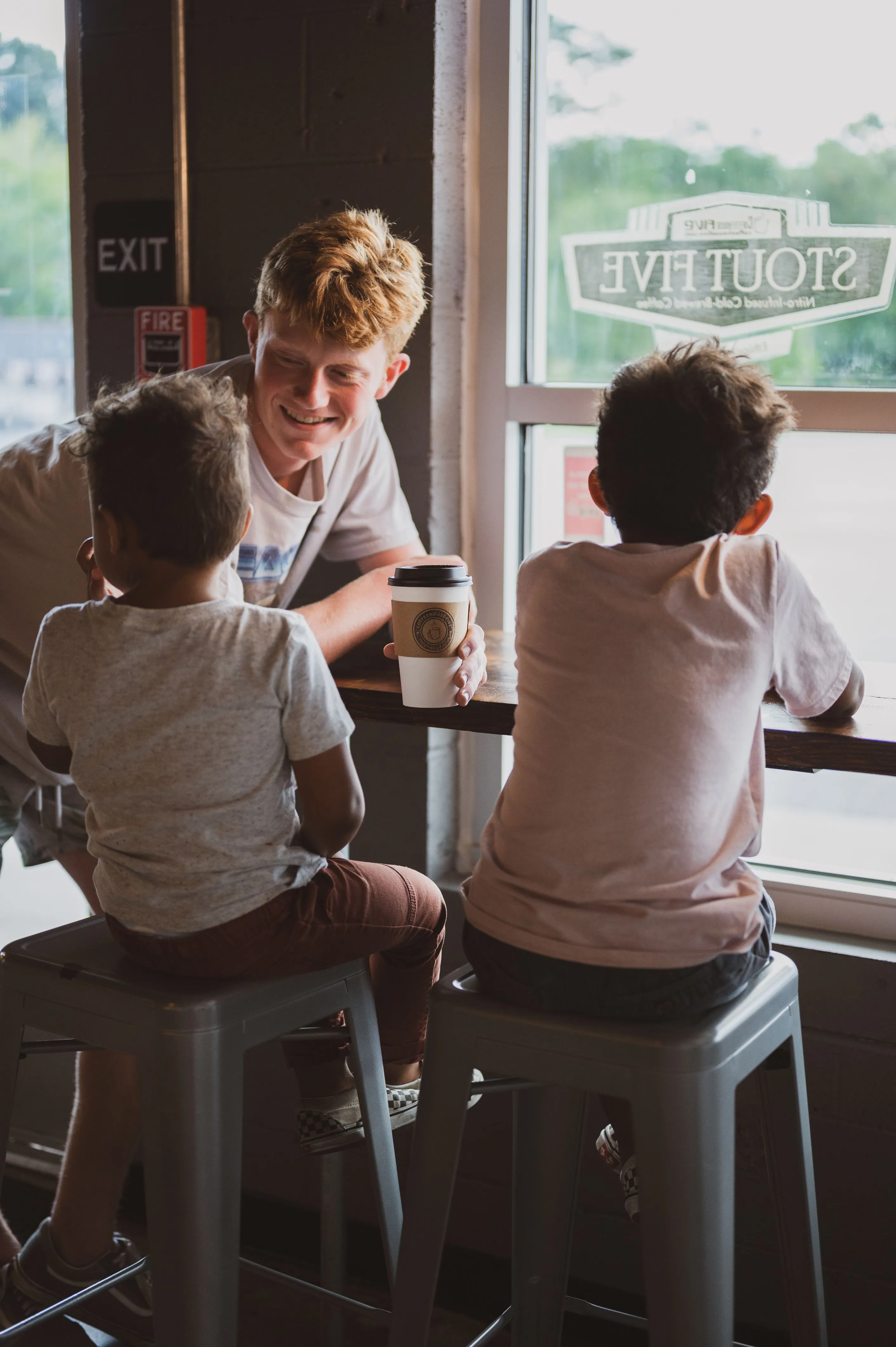 A smiling person sitting on a stool at a café bar next to two children facing a window with a "Southrive" sign, holding a takeout coffee cup.