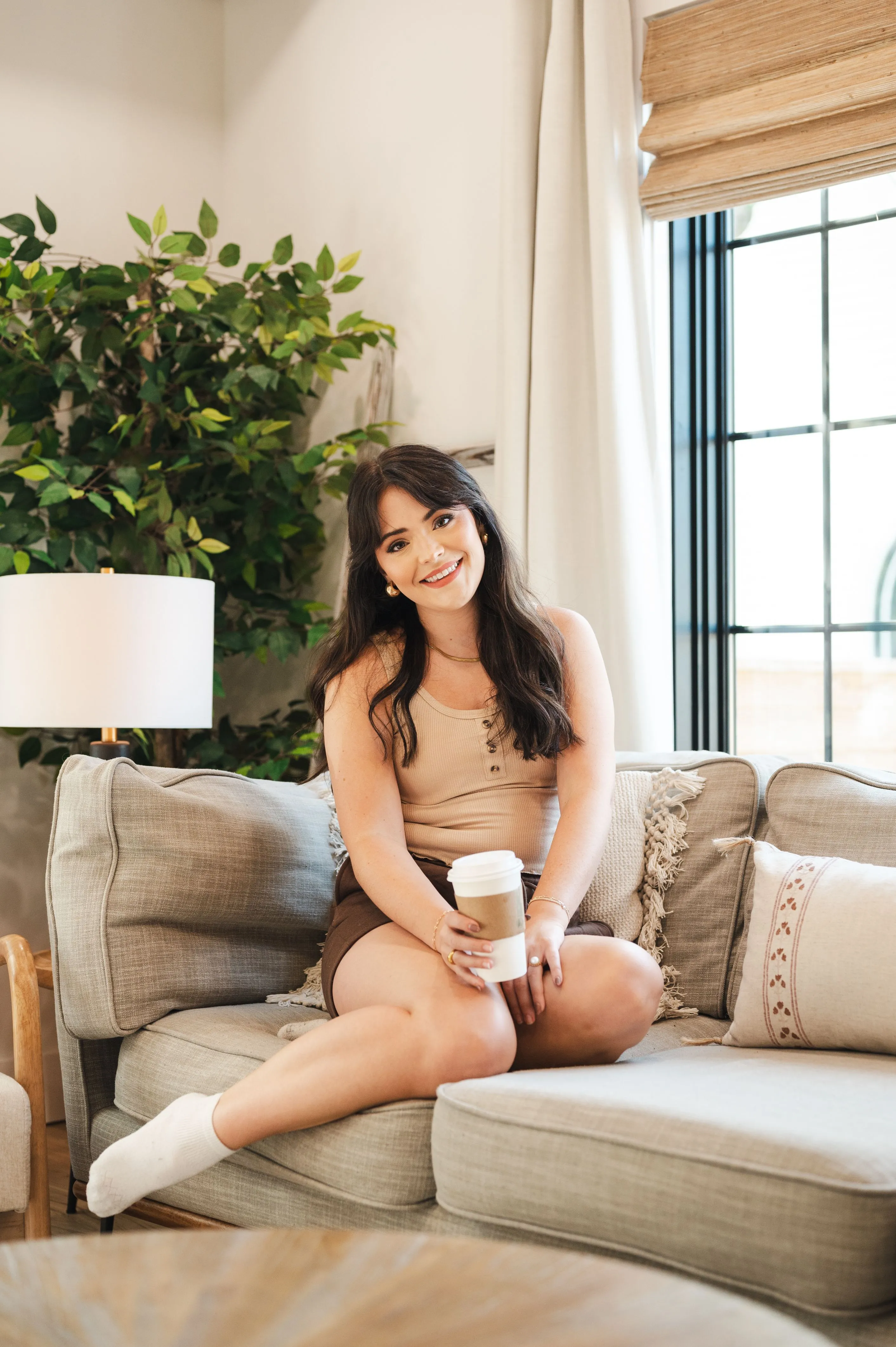 Woman smiling on a sofa holding a coffee cup with indoor plants and natural light in the background.