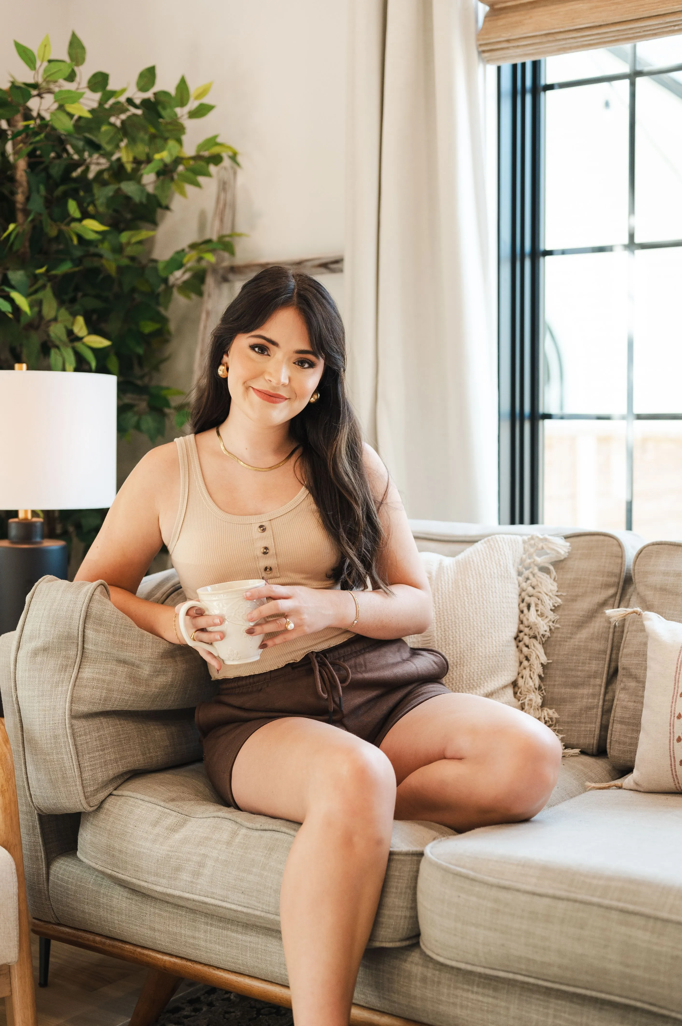Woman smiling while sitting on a couch holding a cup, with a plant and sunlight coming through a window in the background.