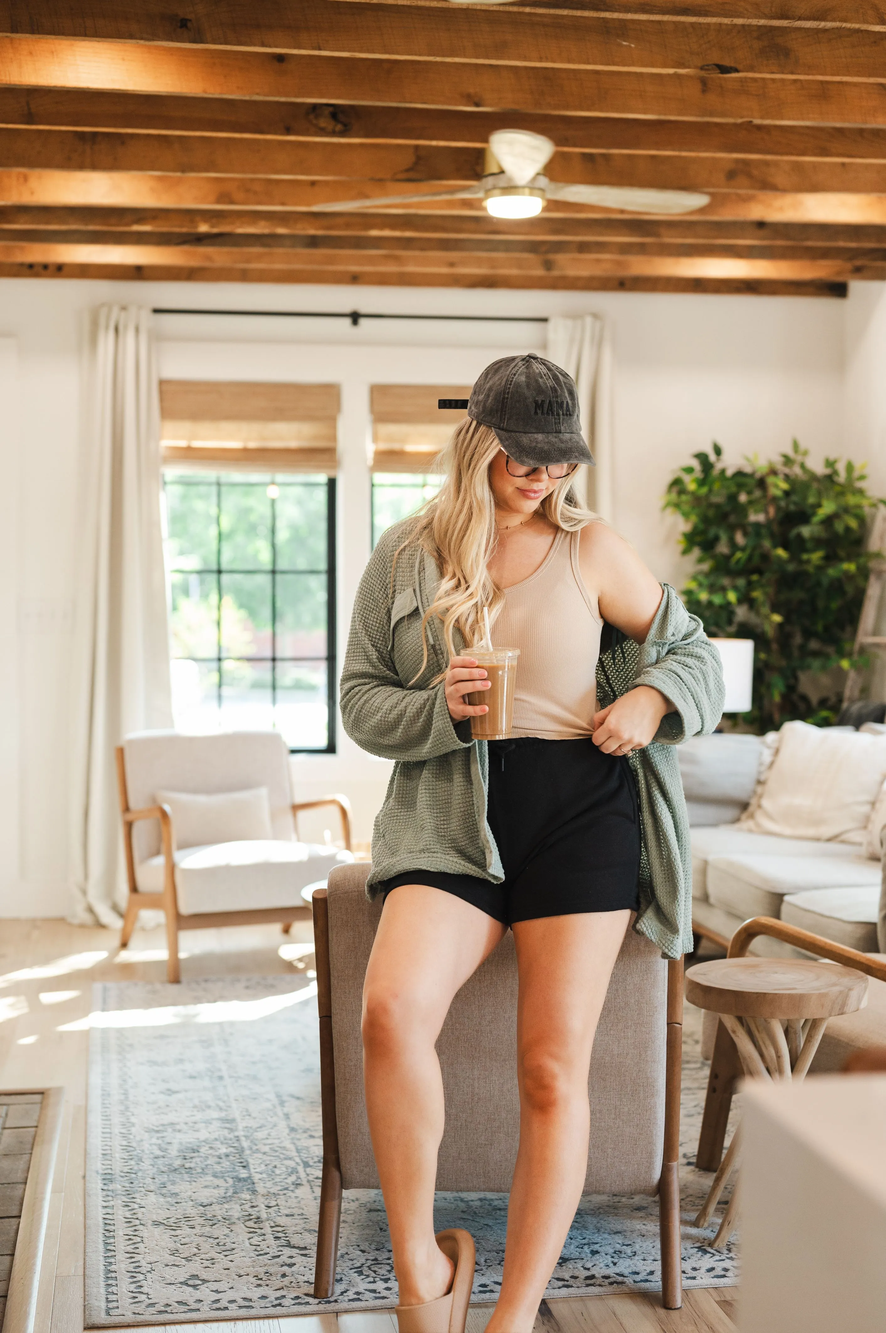 Woman in a casual outfit with a cap, holding a coffee cup, standing indoors in a cozy living room with wooden accents.