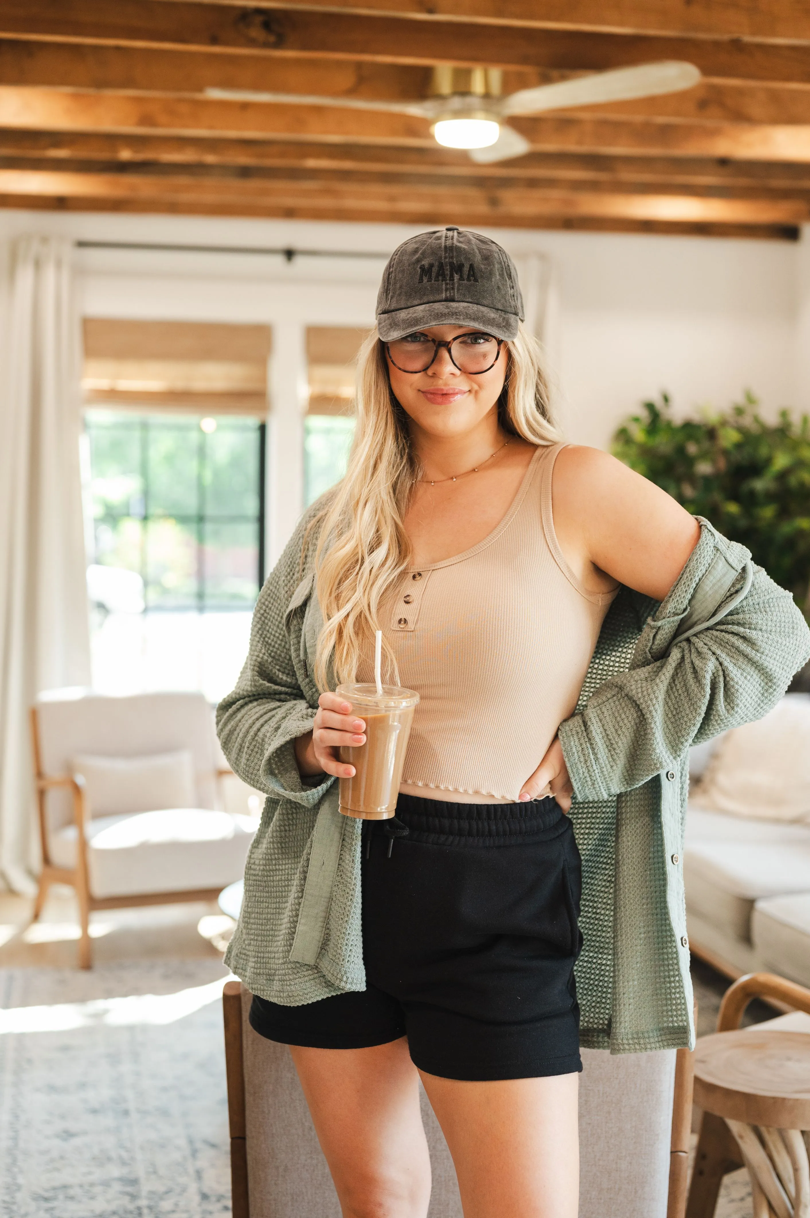 Smiling woman standing indoors wearing a beige tank top, black shorts, green cardigan, and a gray "MAMA" hat, holding an iced coffee drink.