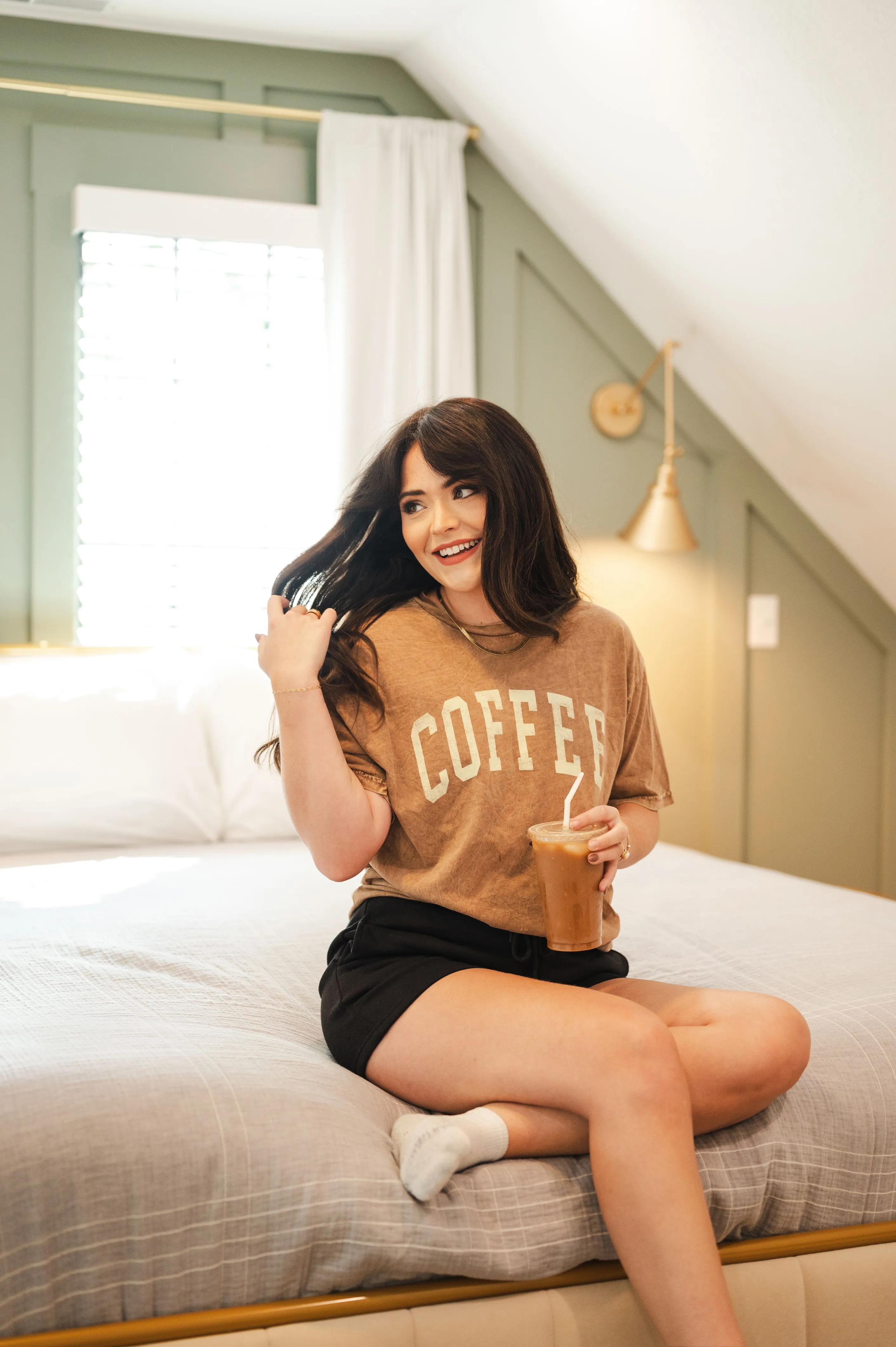 Young woman sitting on a bed holding an iced coffee and smiling, with a t-shirt that reads "COFFEE" in a brightly lit room.