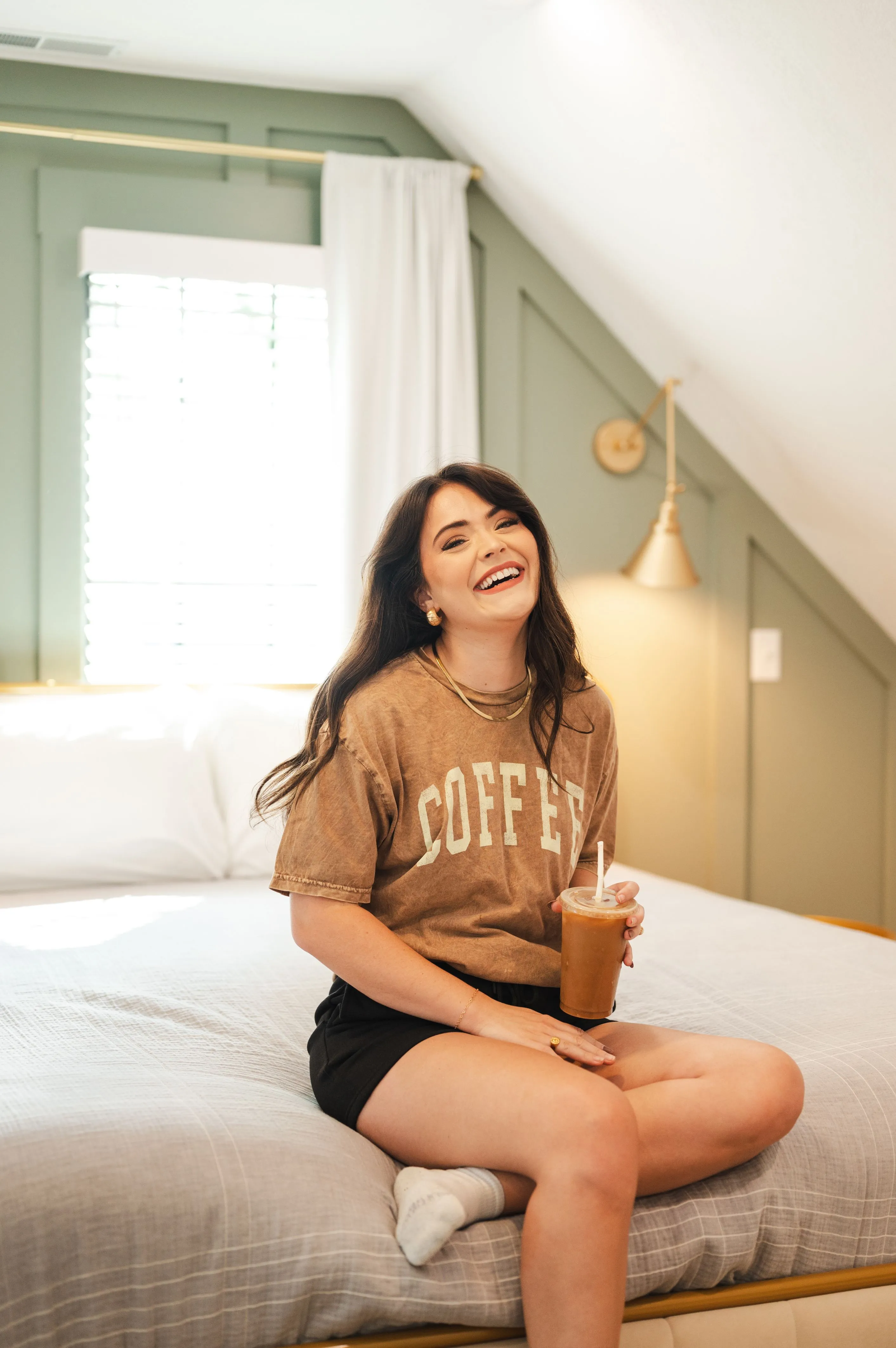 Woman sitting on a bed, laughing, holding an iced coffee, wearing a brown T-shirt with the word "COFFEE" on it and black shorts, in a room with green walls and white curtains.
