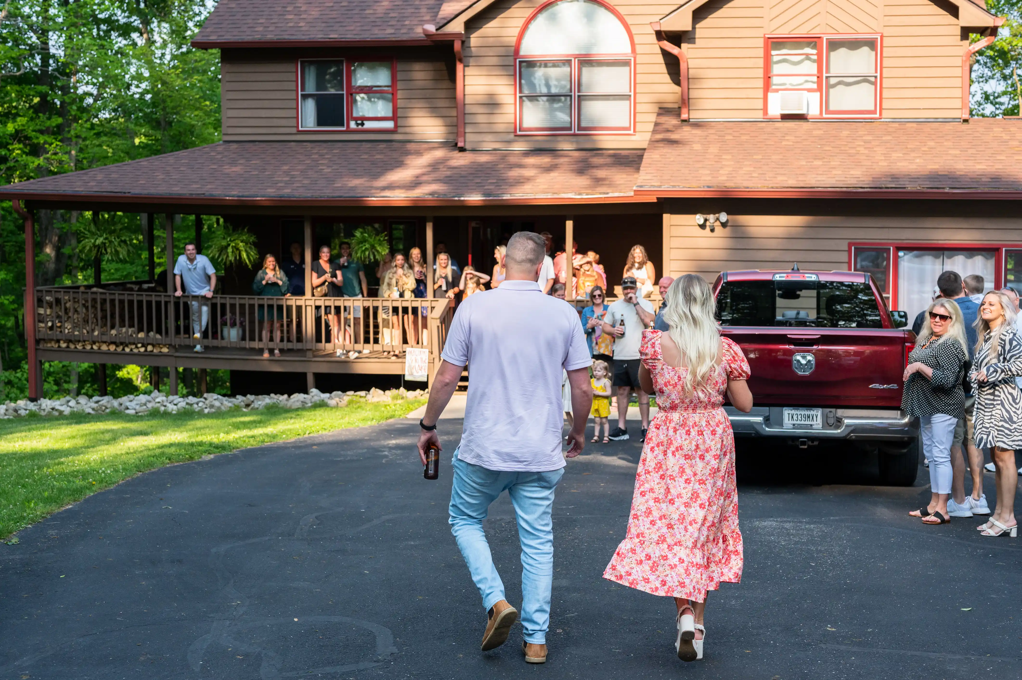 Two people walking towards a house where a group of guests are gathered on the porch for a social event.