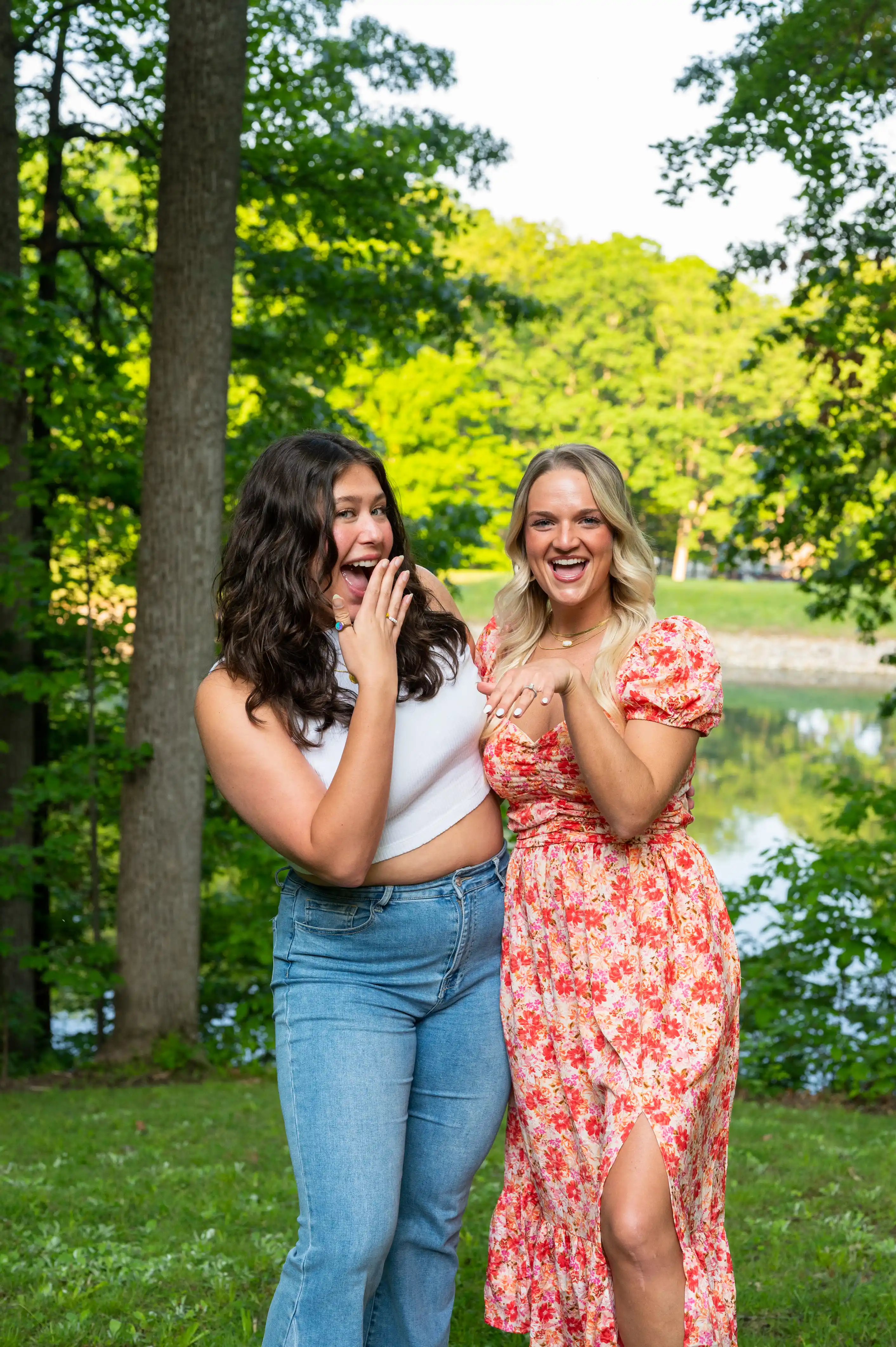 Two smiling women standing outdoors, one gesturing a shush sign, with trees and a pond in the background.