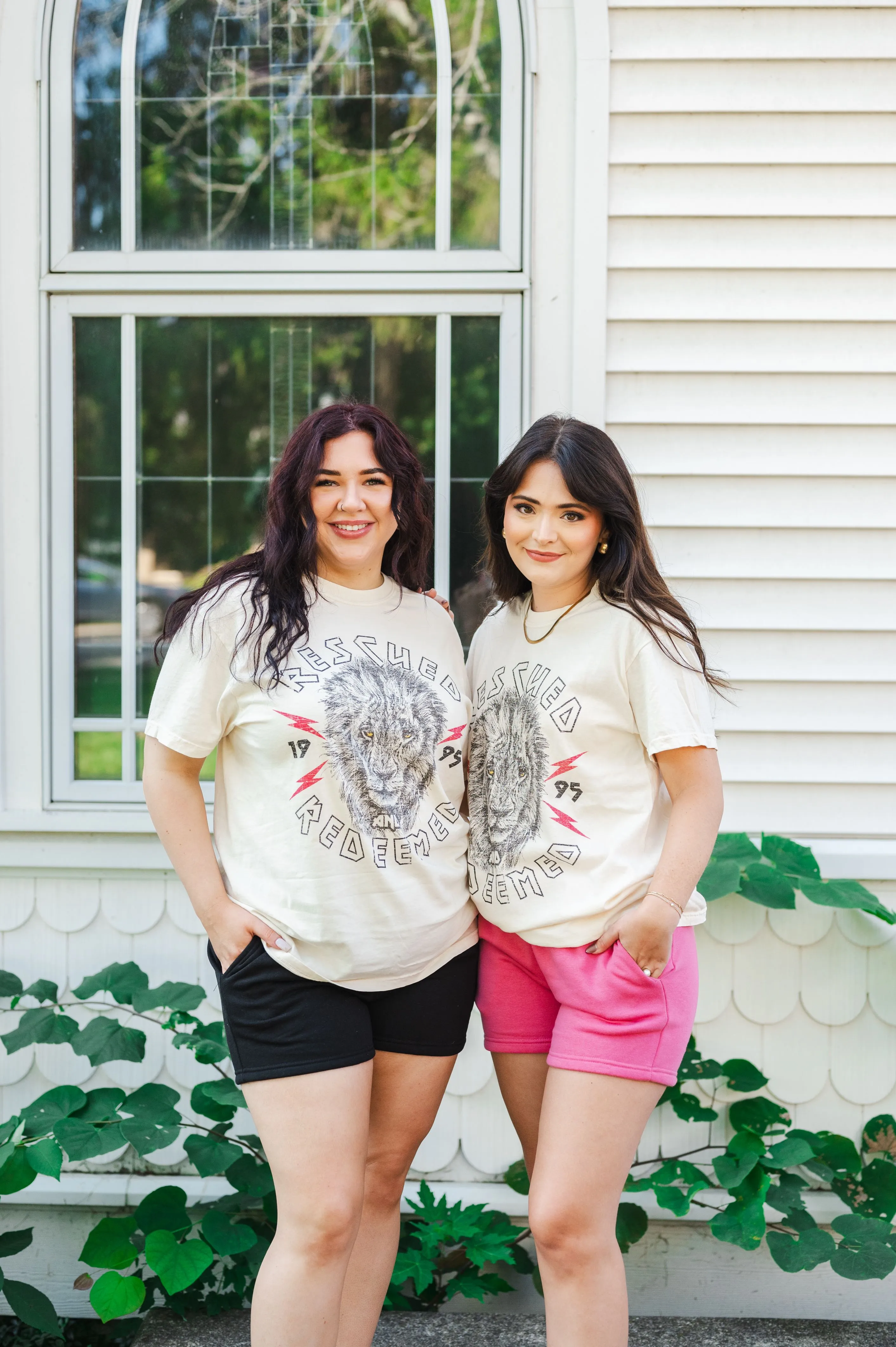 Two smiling women standing together in front of a house with a white window, wearing matching beige T-shirts with a wolf print design and different colored shorts.