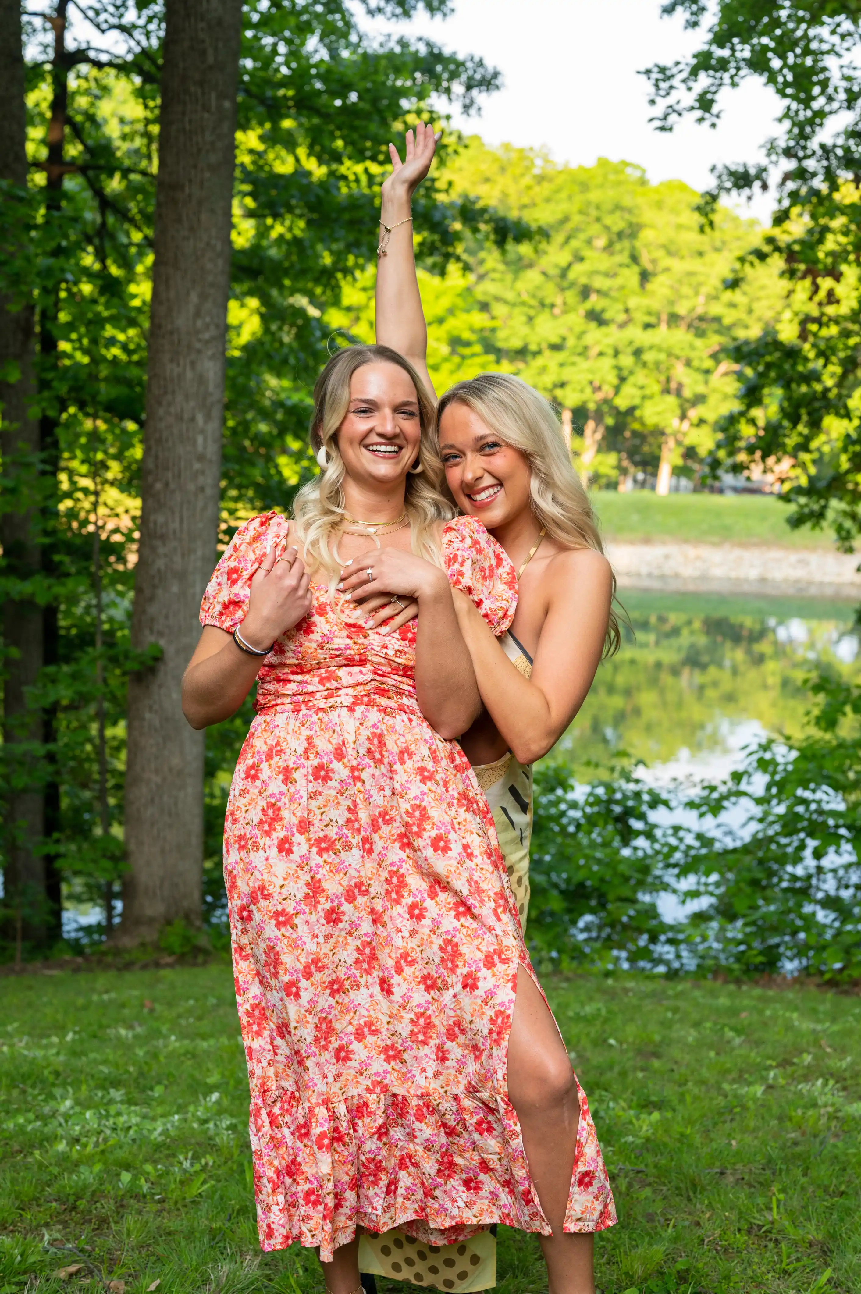 Two women smiling and hugging each other in a park with trees and a pond in the background. They are both wearing floral dresses.