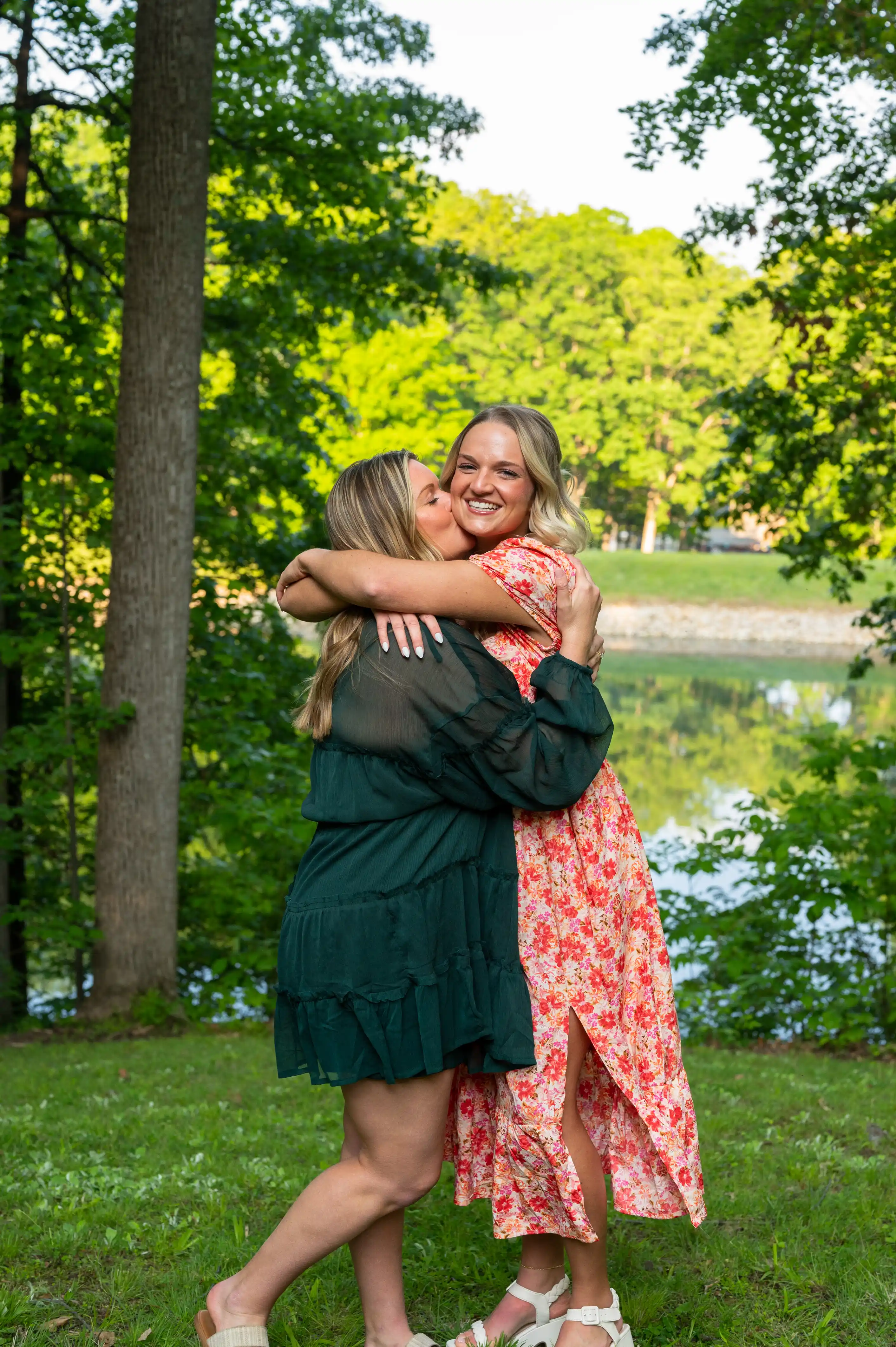 Two women embracing in a joyful hug outdoors with trees and a lake in the background.