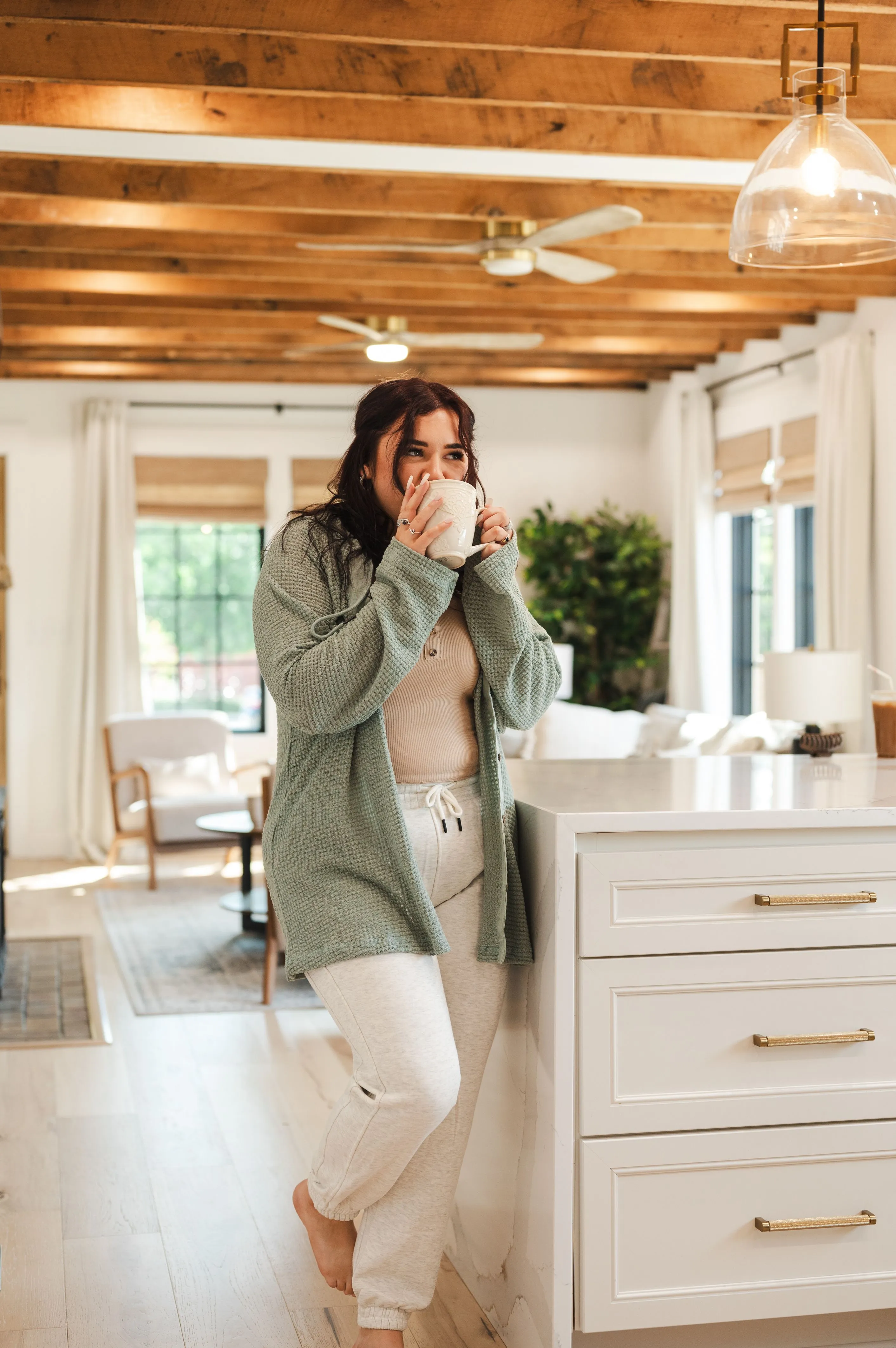 Woman in cozy attire leaning on kitchen counter sipping from a mug.