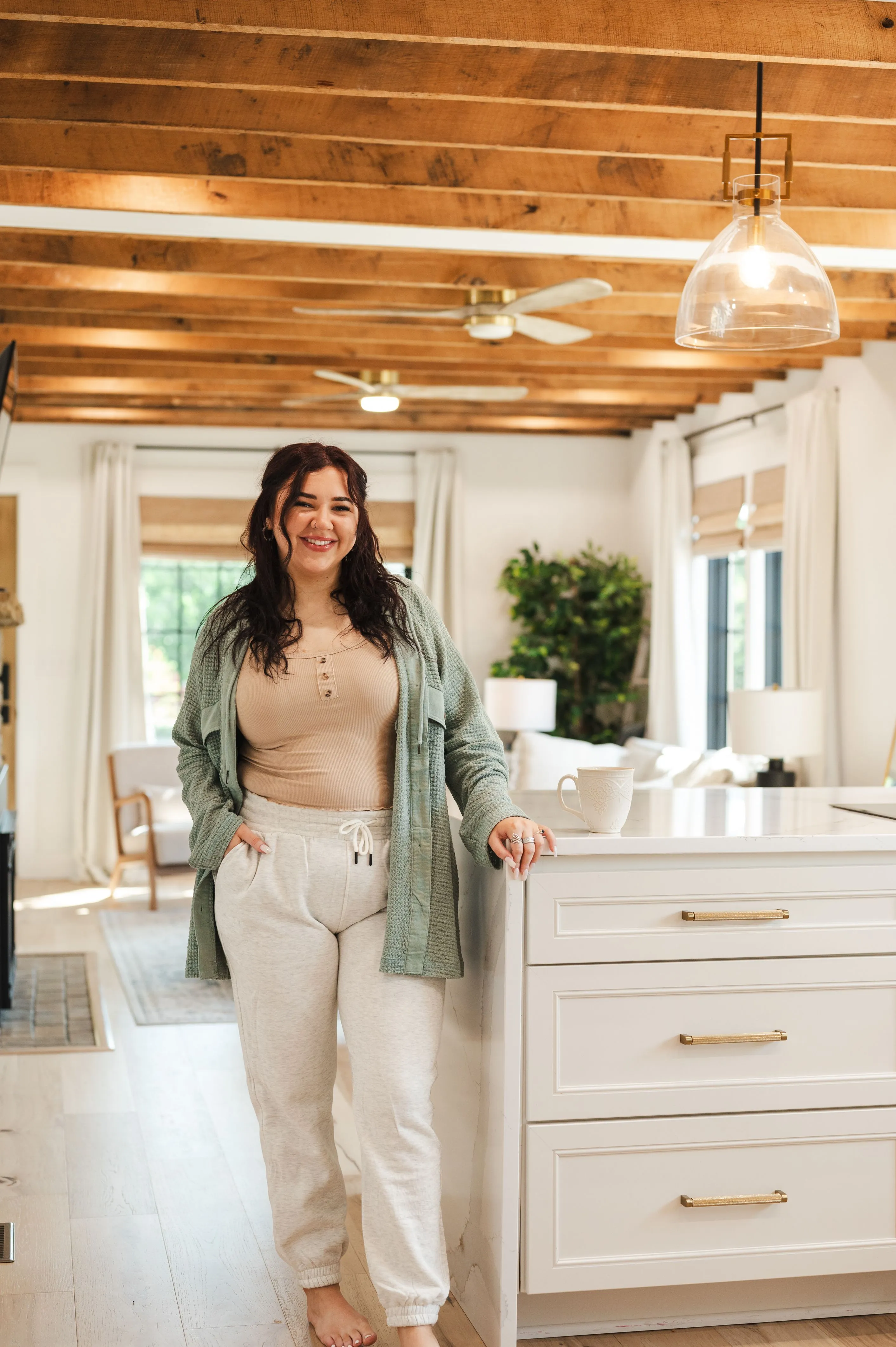 Woman smiling in a cozy home interior with exposed wooden ceiling beams, leaning on a kitchen island next to a white mug.
