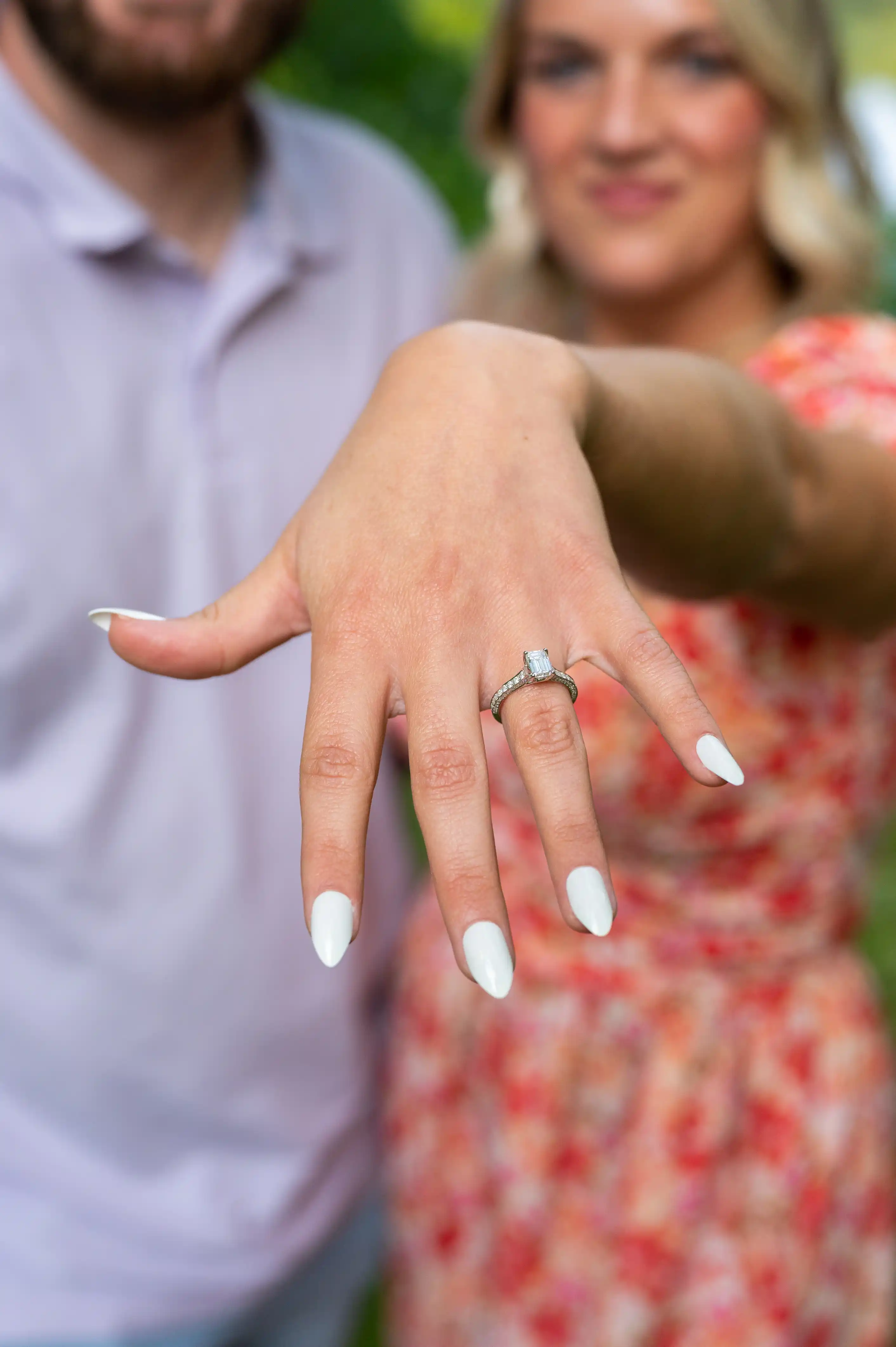 Woman showing engagement ring on her finger with an out-of-focus man in the background.
