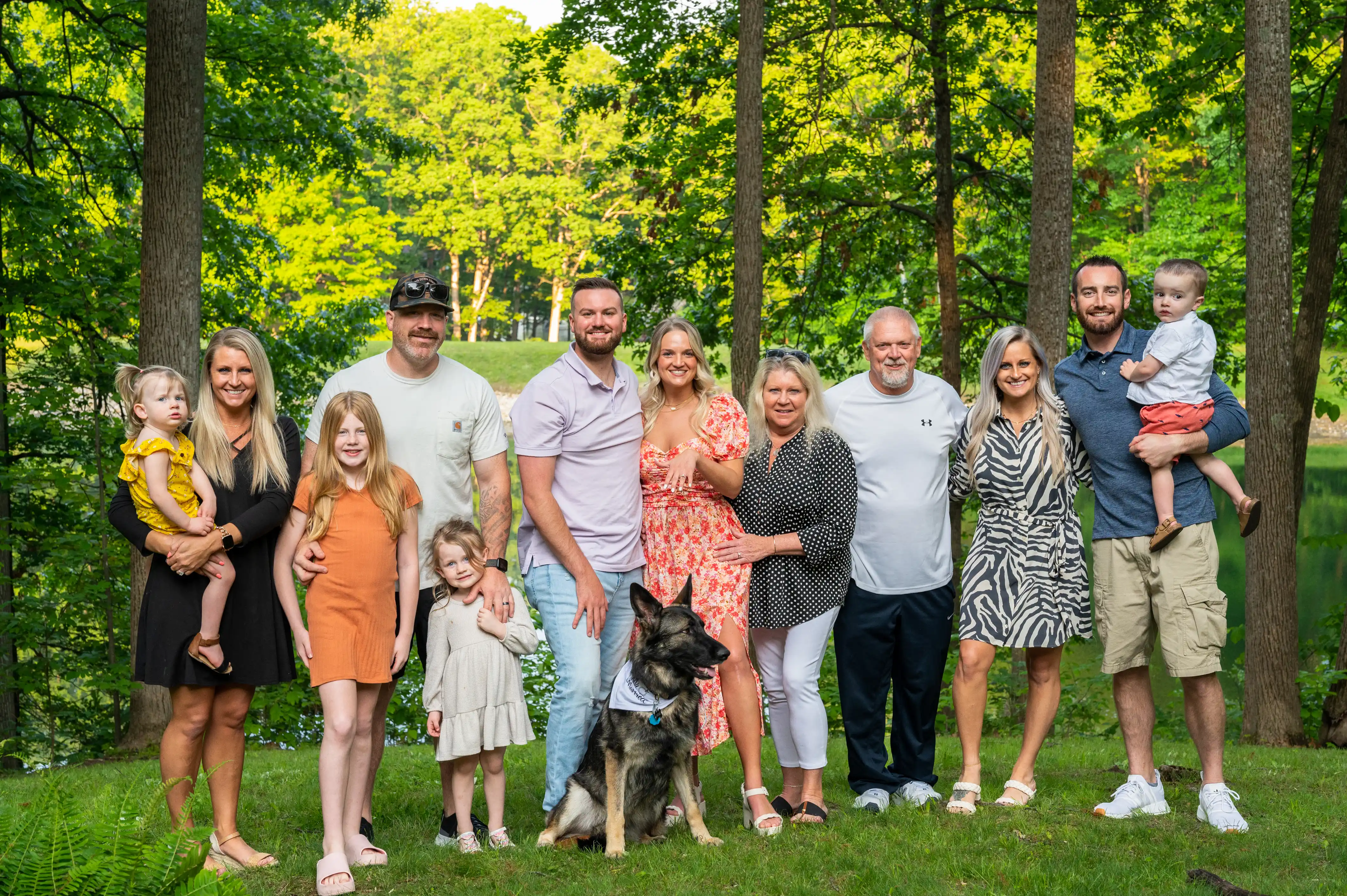 A happy family with multiple generations smiles for a photo outdoors, with trees in the background. A dog is also included in the family portrait.
