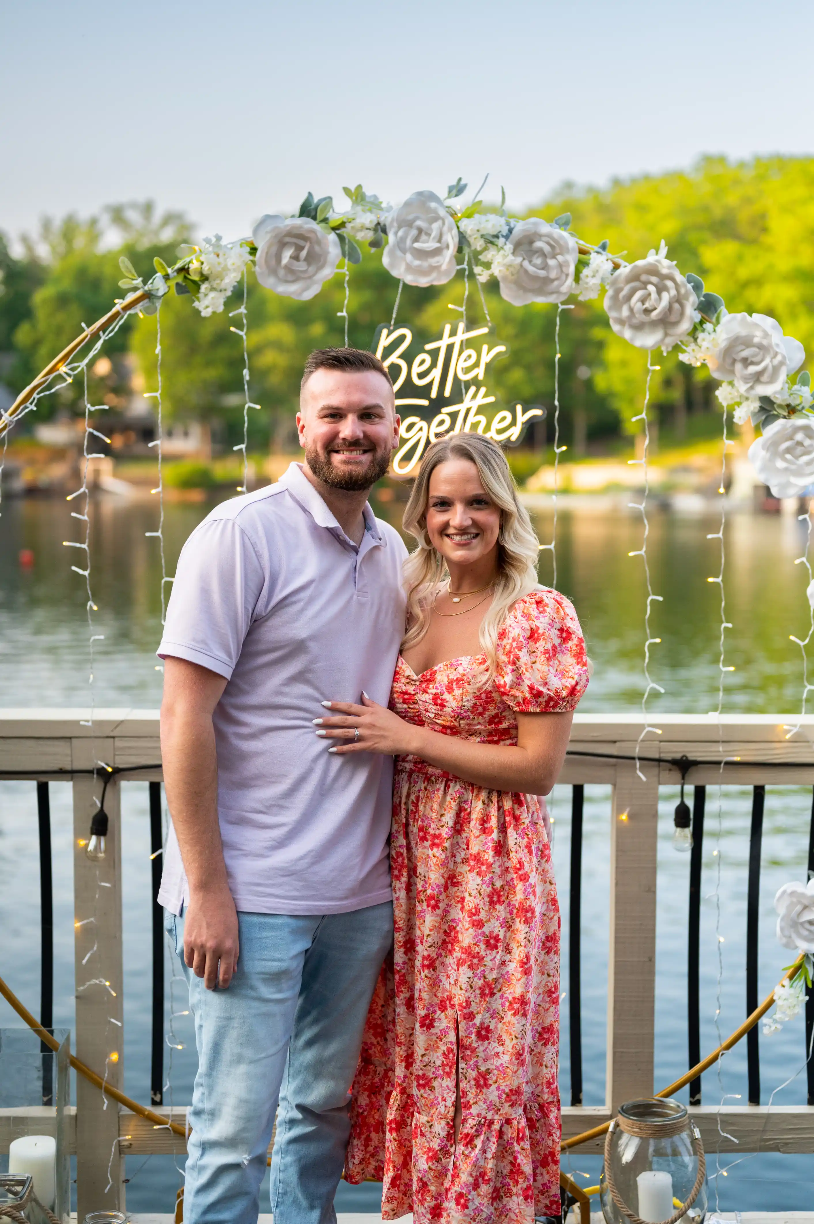 Smiling couple posing for a photo at a baby shower event with a lake and trees in the background, decorations with "Baby Shower" text overhead.