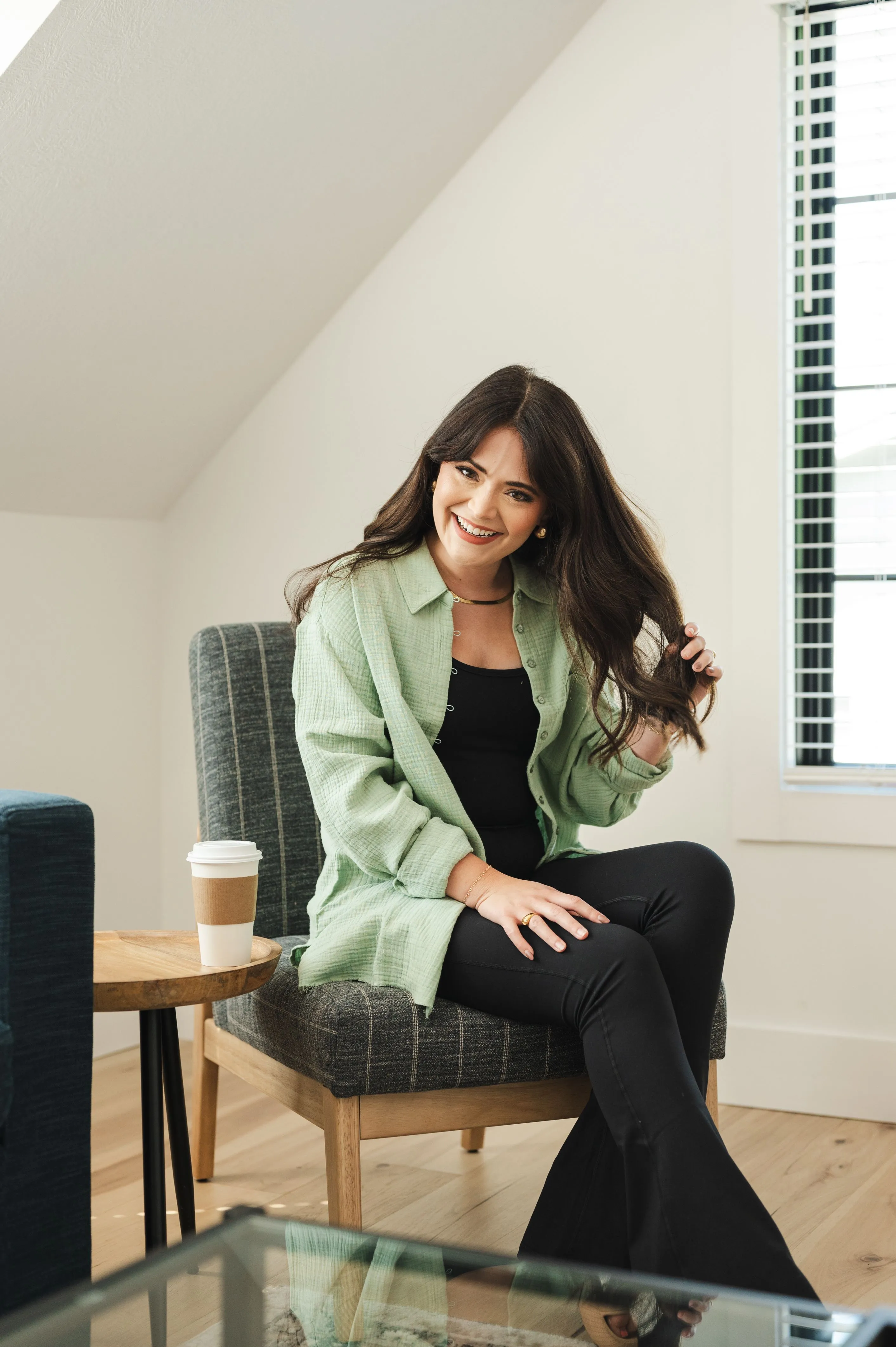 Smiling woman sitting casually on a chair in a well-lit room with a coffee cup on a side table.
