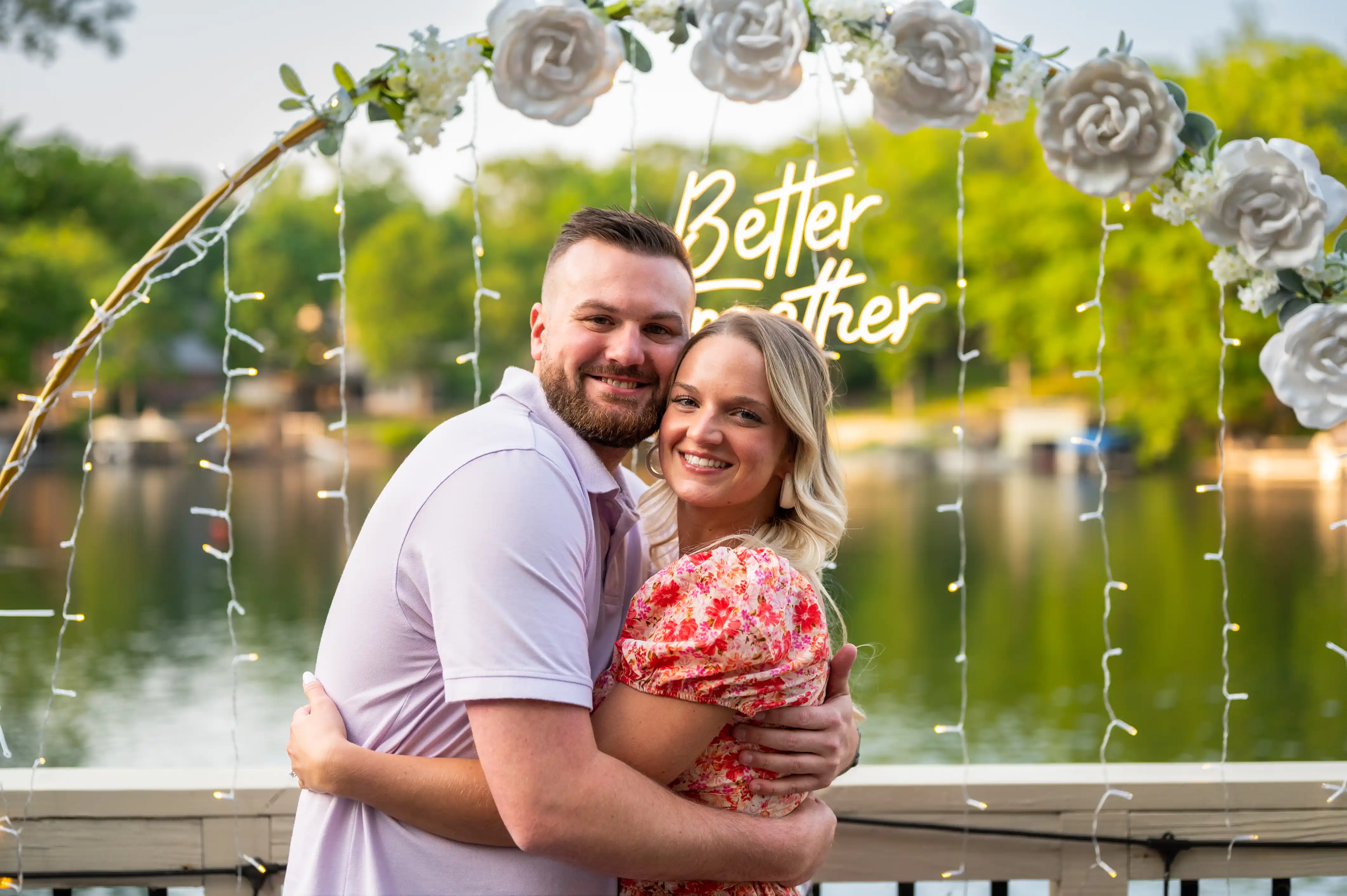 Couple embracing near a lake with the phrase "Better Together" displayed above them in decorative writing, surrounded by a floral design.