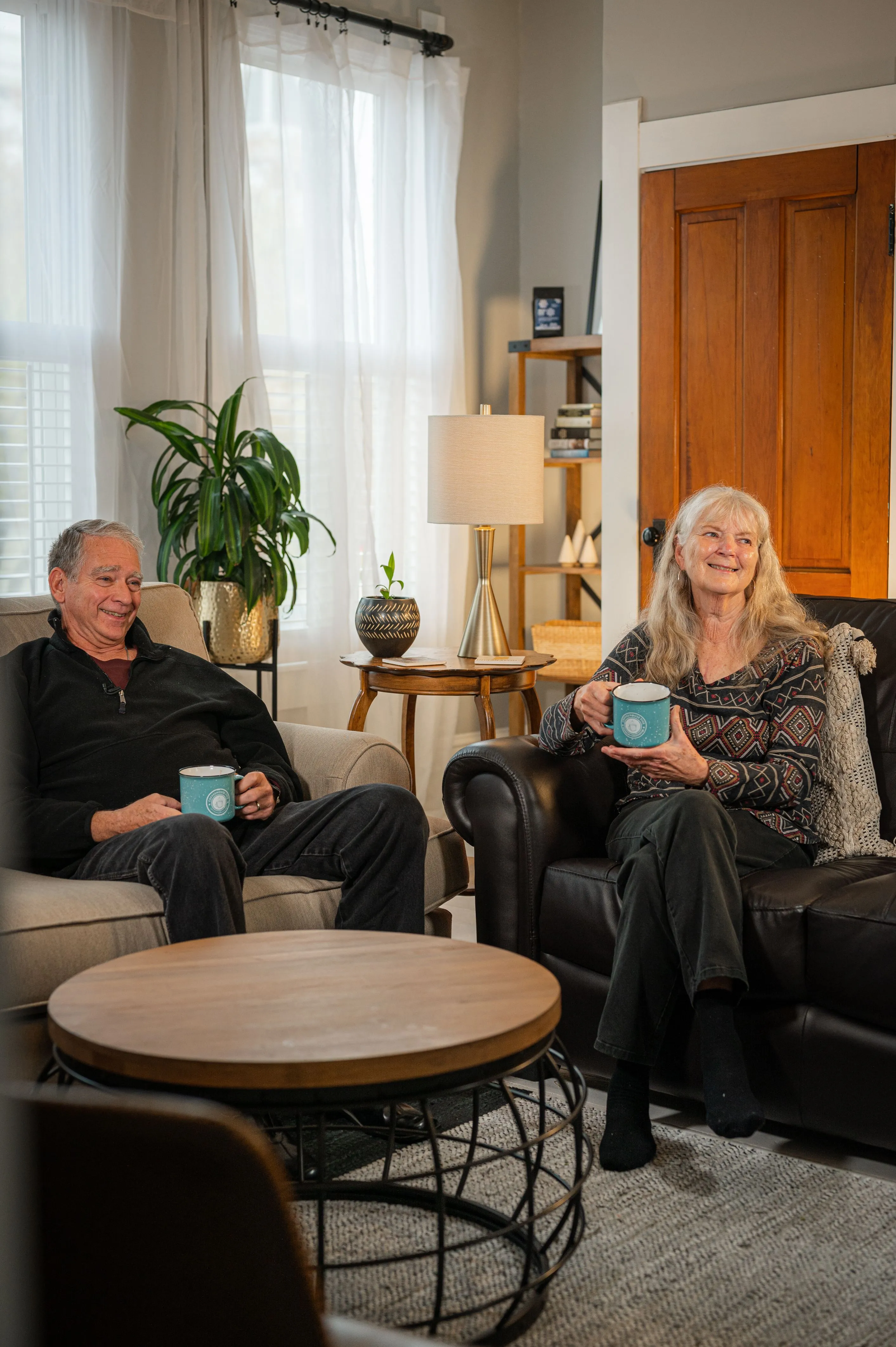 An elderly couple smiling and relaxing in a cozy living room with mugs in their hands.