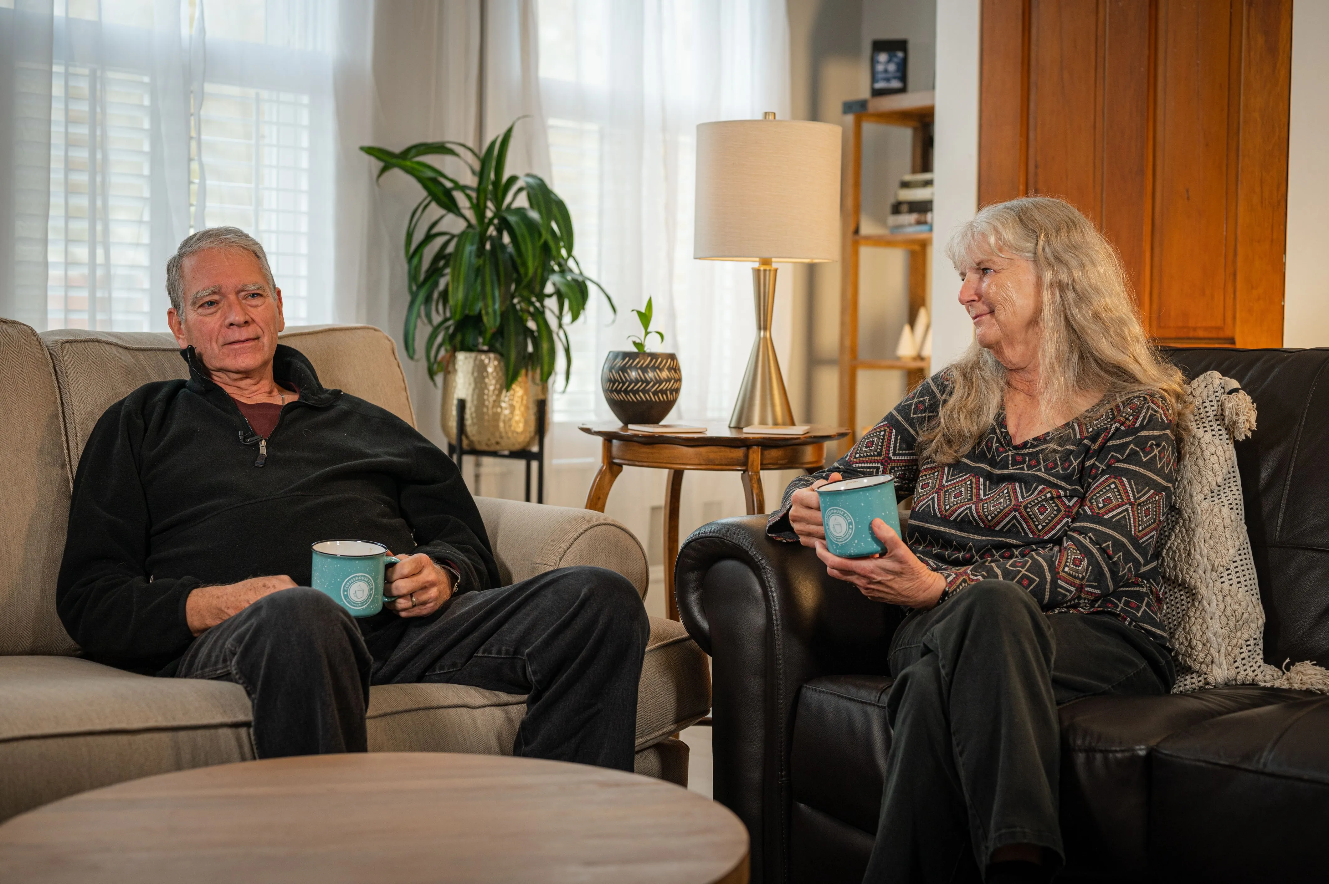 Two senior adults relax on a sofa while holding mugs in a cozy living room with a plant and lamp in the background.