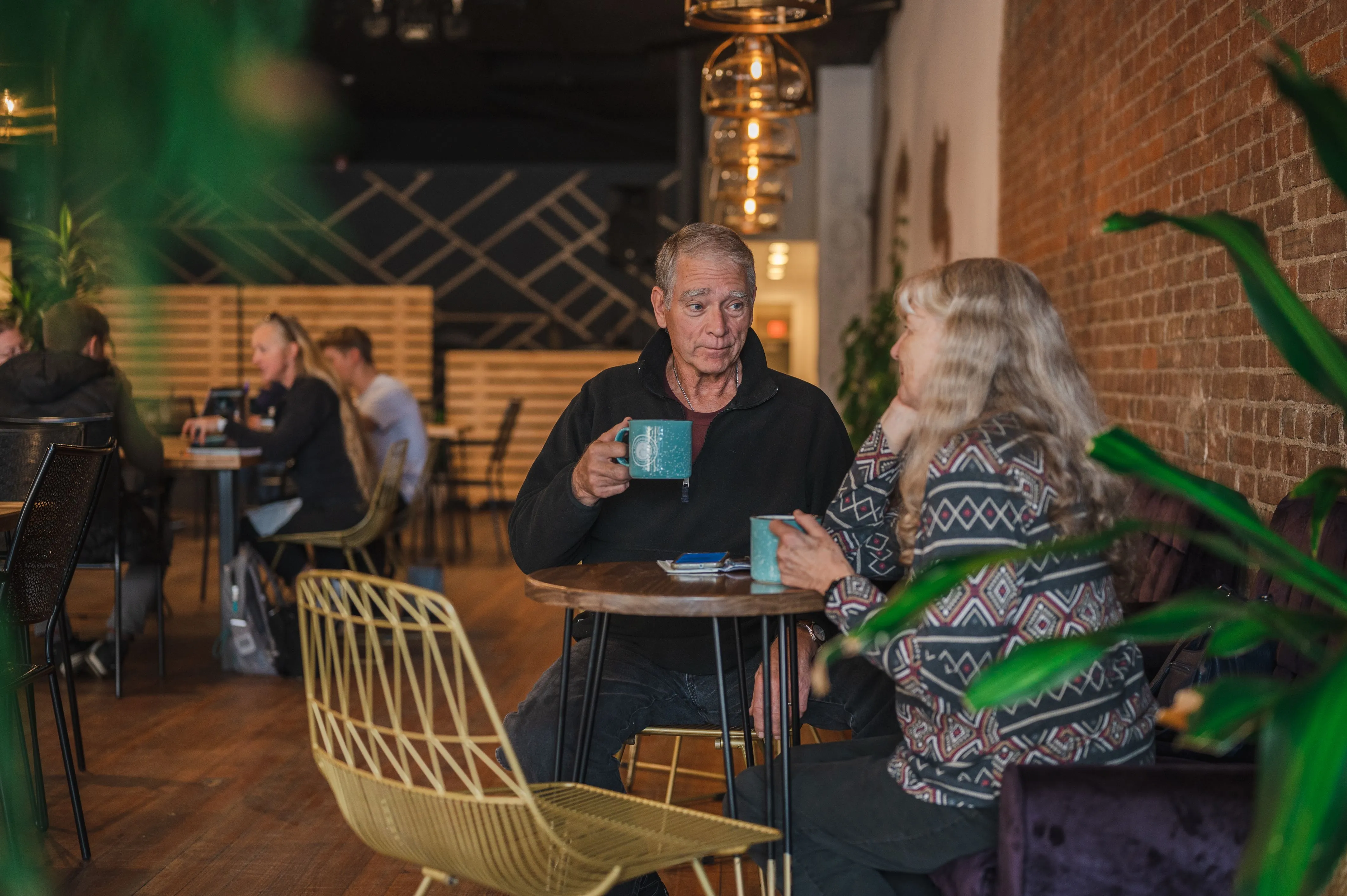 Two people, a man and a woman, having a conversation over coffee in a cozy cafe with other patrons and brick walls in the background.