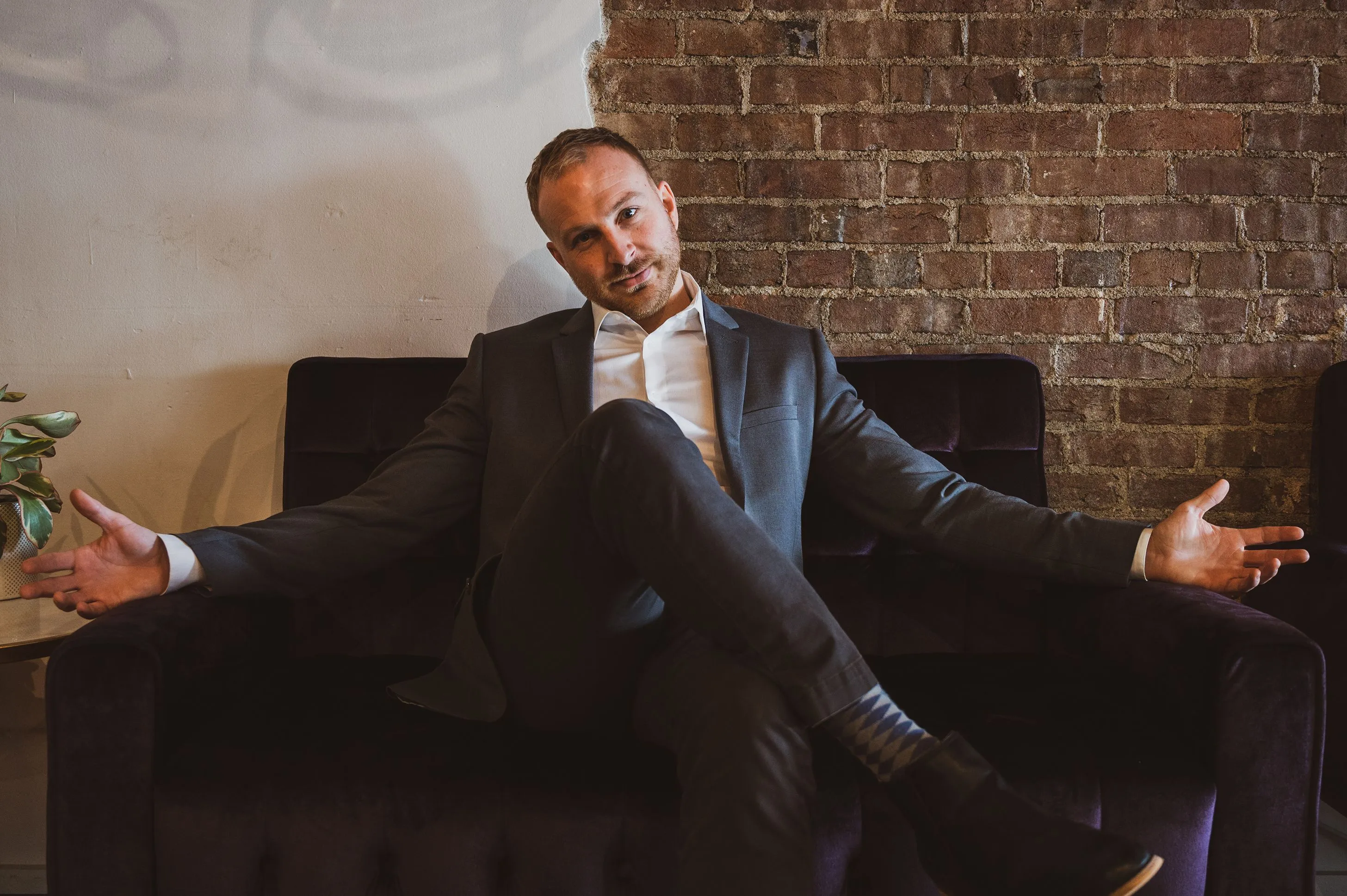 Man in a suit sitting relaxed with arms outstretched on a couch against a brick wall background.
