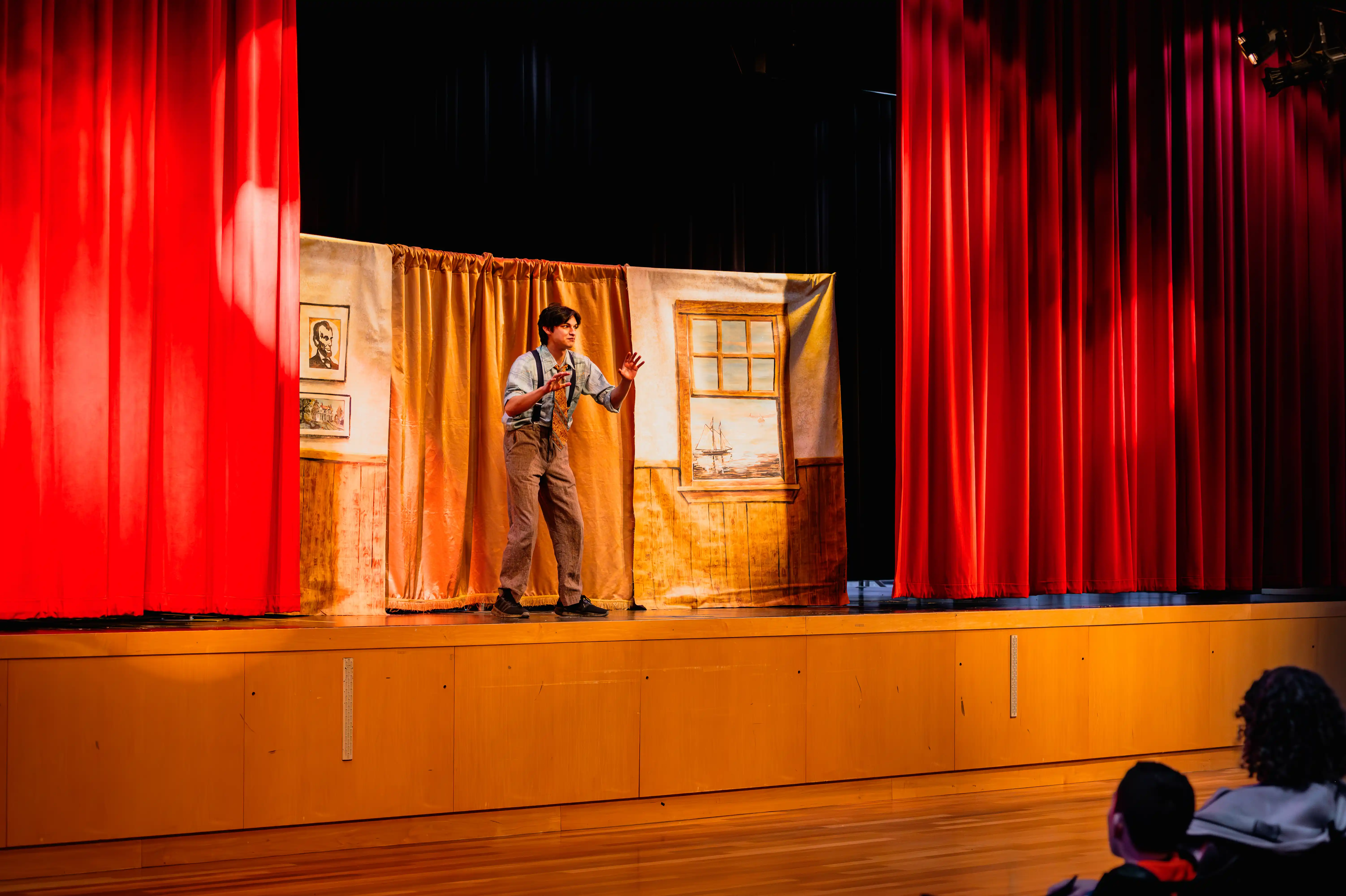 Performer on stage at a theater with red curtains and a simple set, addressing the audience.