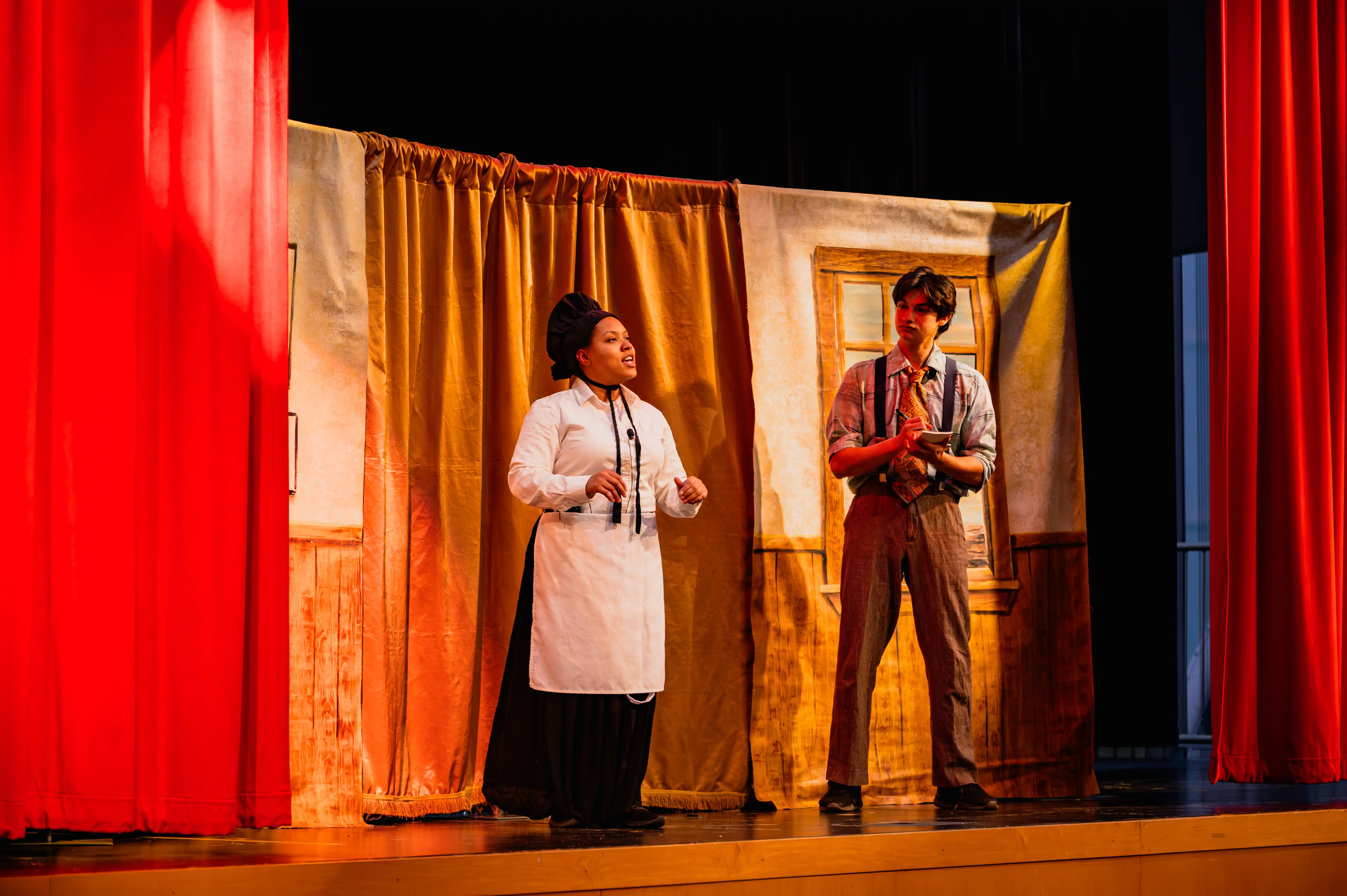 Two actors on stage performing in a play, with one dressed as a chef and the other wearing suspenders, in front of a curtain.