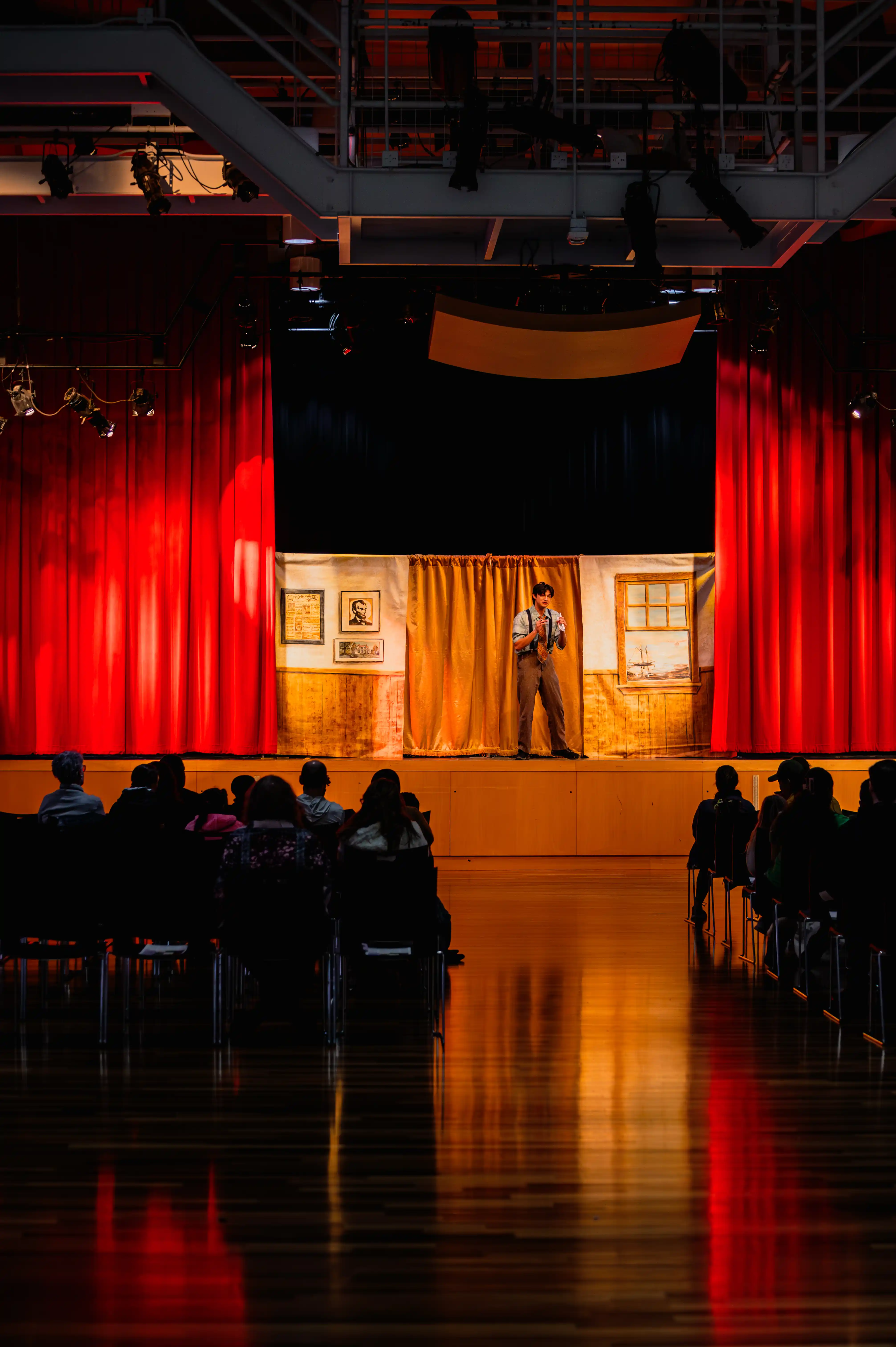 Audience seated in dimmed light watching a lone performer on a brightly lit stage framed by red curtains.