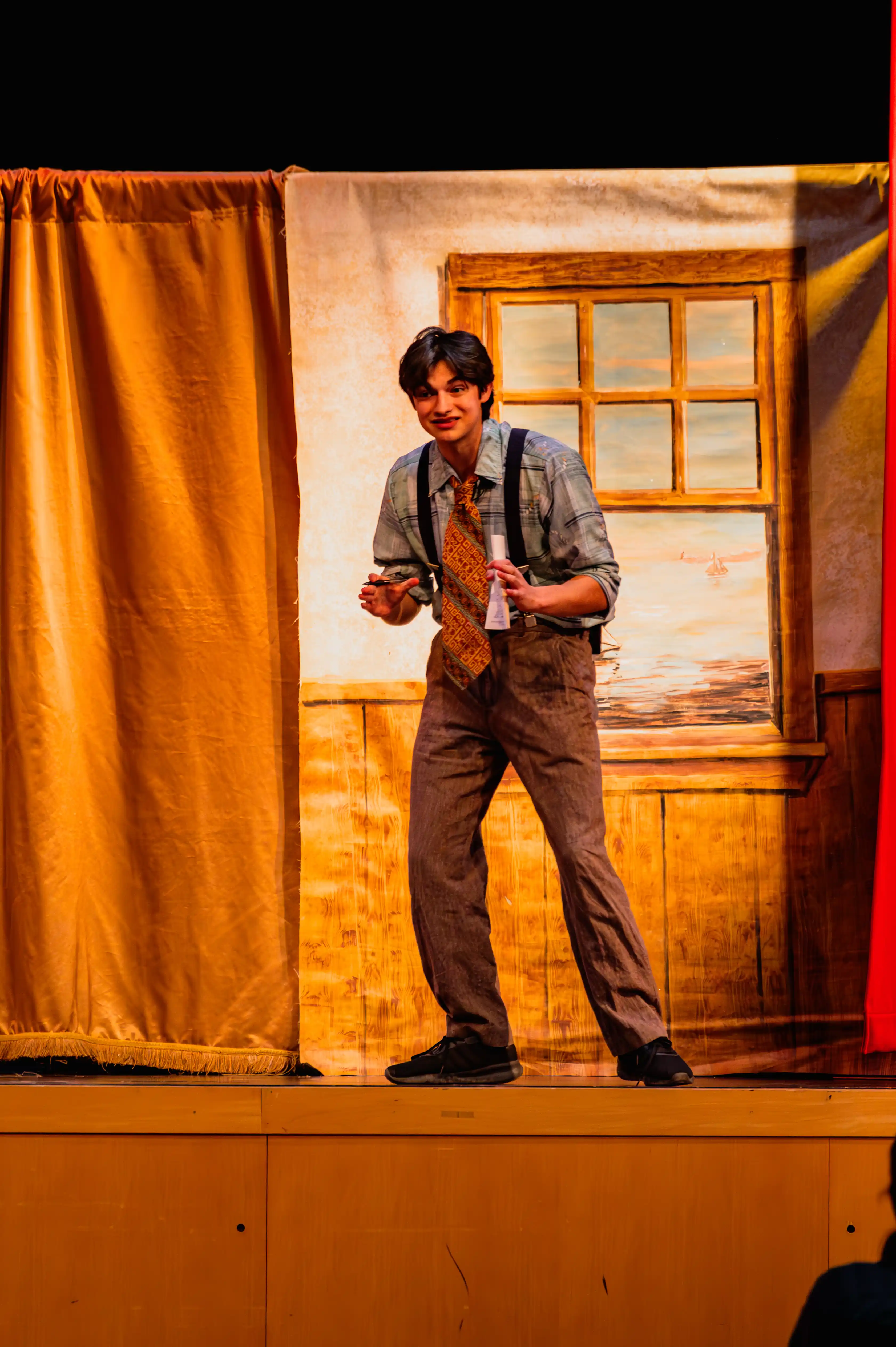 Actor on stage performing a scene with expressive body language, dressed in vintage attire with suspenders and tie, framed by stage curtains against a warmly lit backdrop.