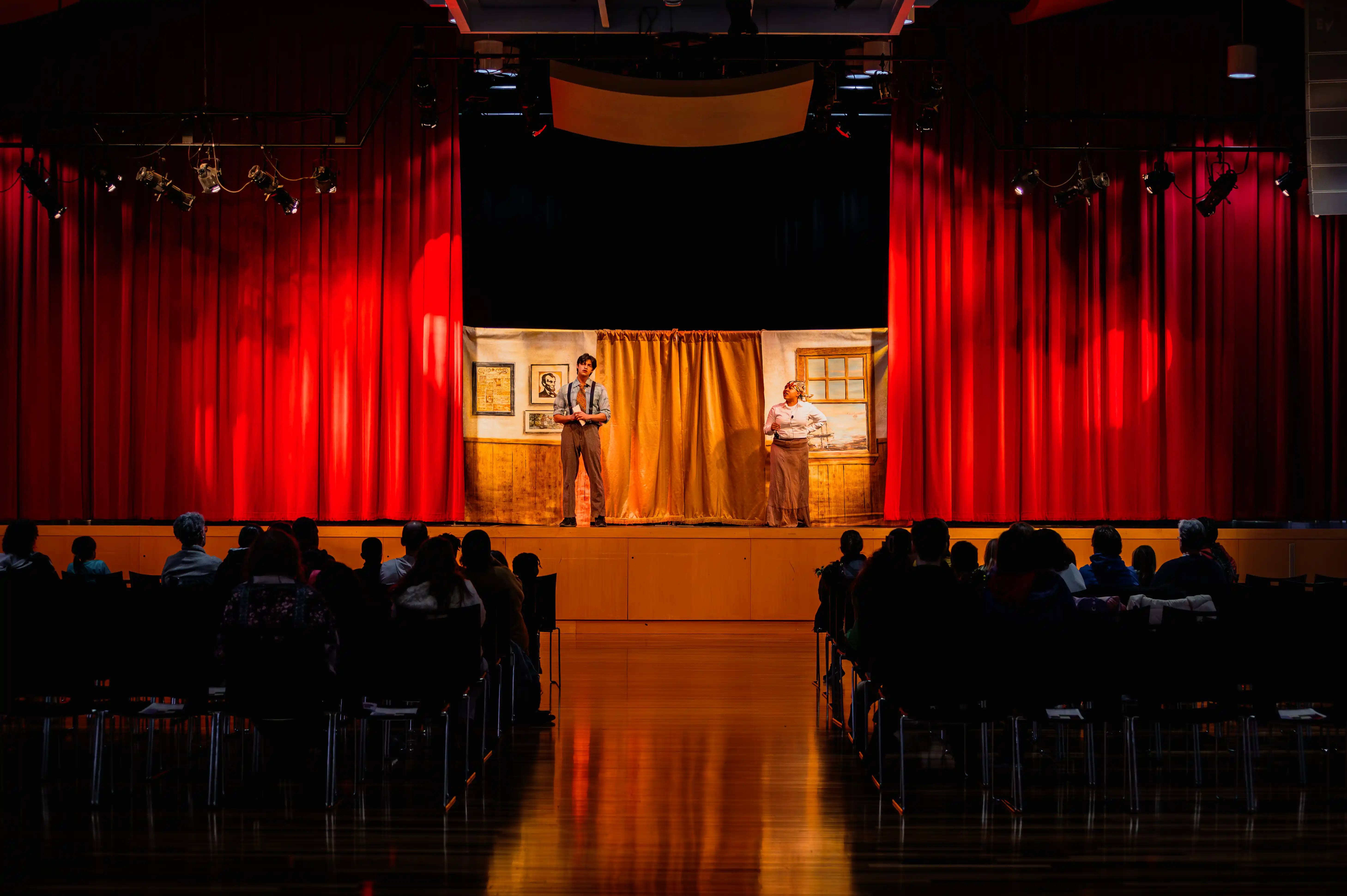 Audience watching two actors on stage in an intimate theater setting with red curtains.