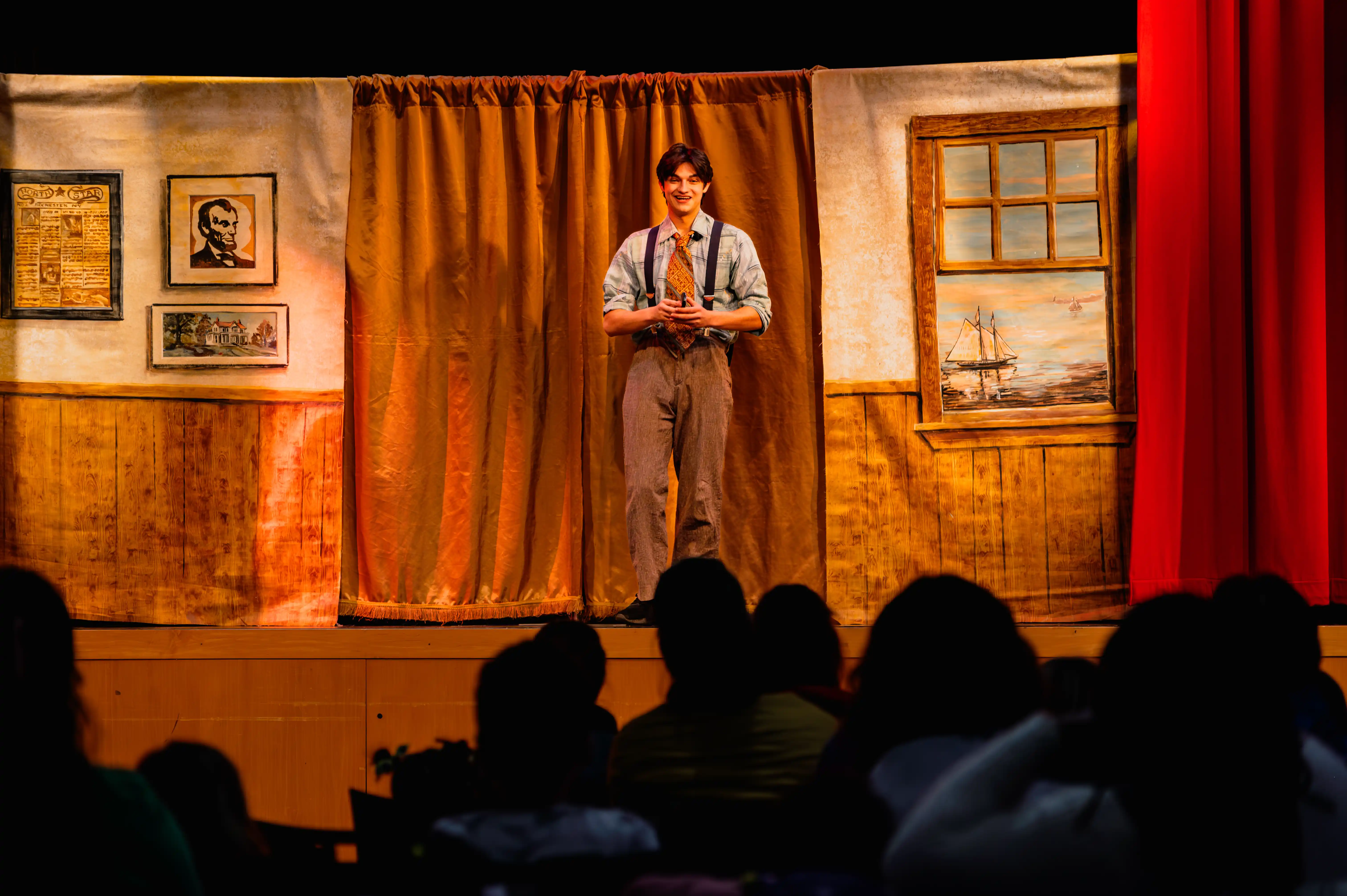 Actor on stage performing in front of an audience with a rustic set design in the background.