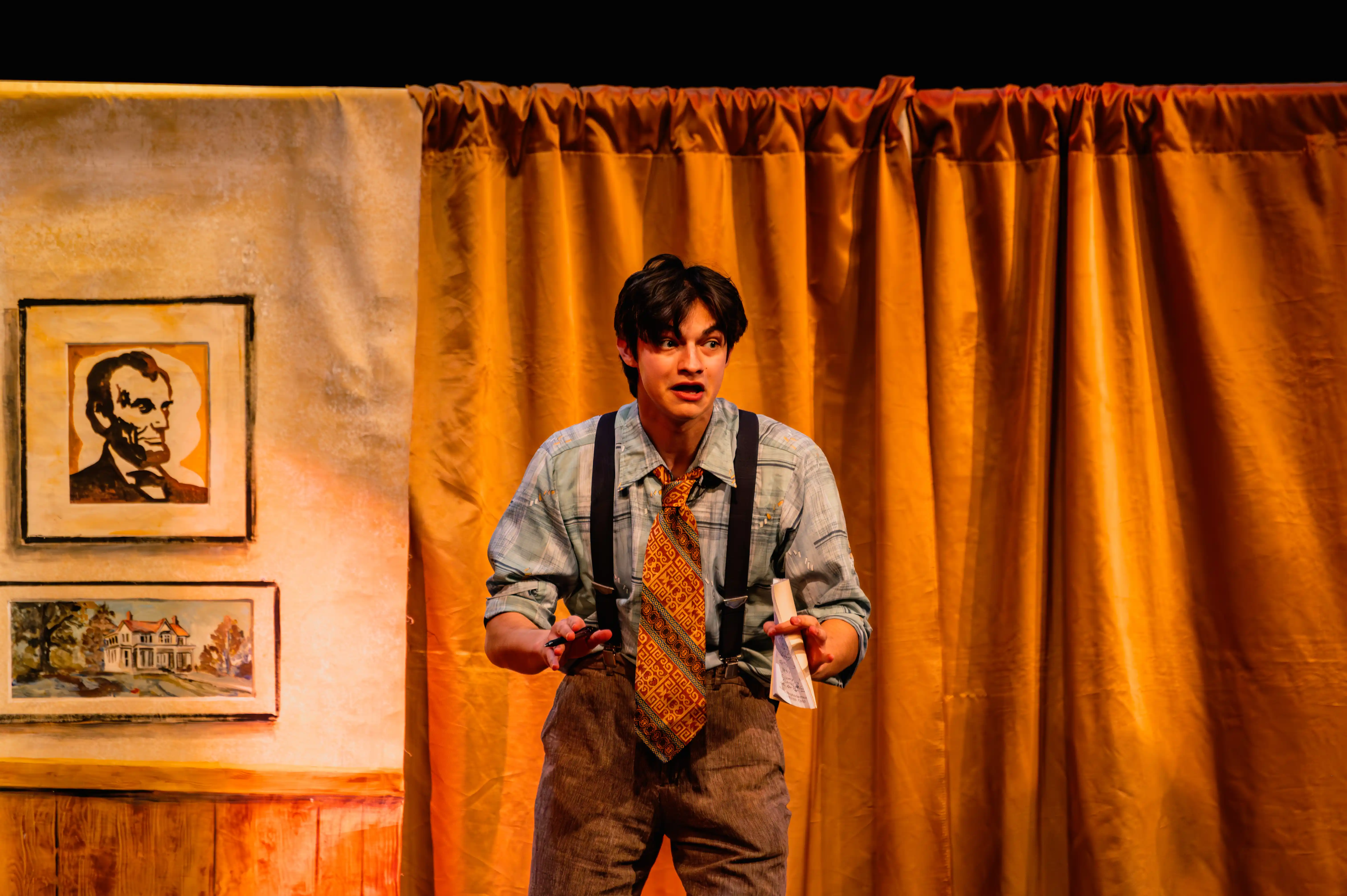 Actor on stage performing with a surprised expression, in front of a yellow curtain with pictures and props in the background.