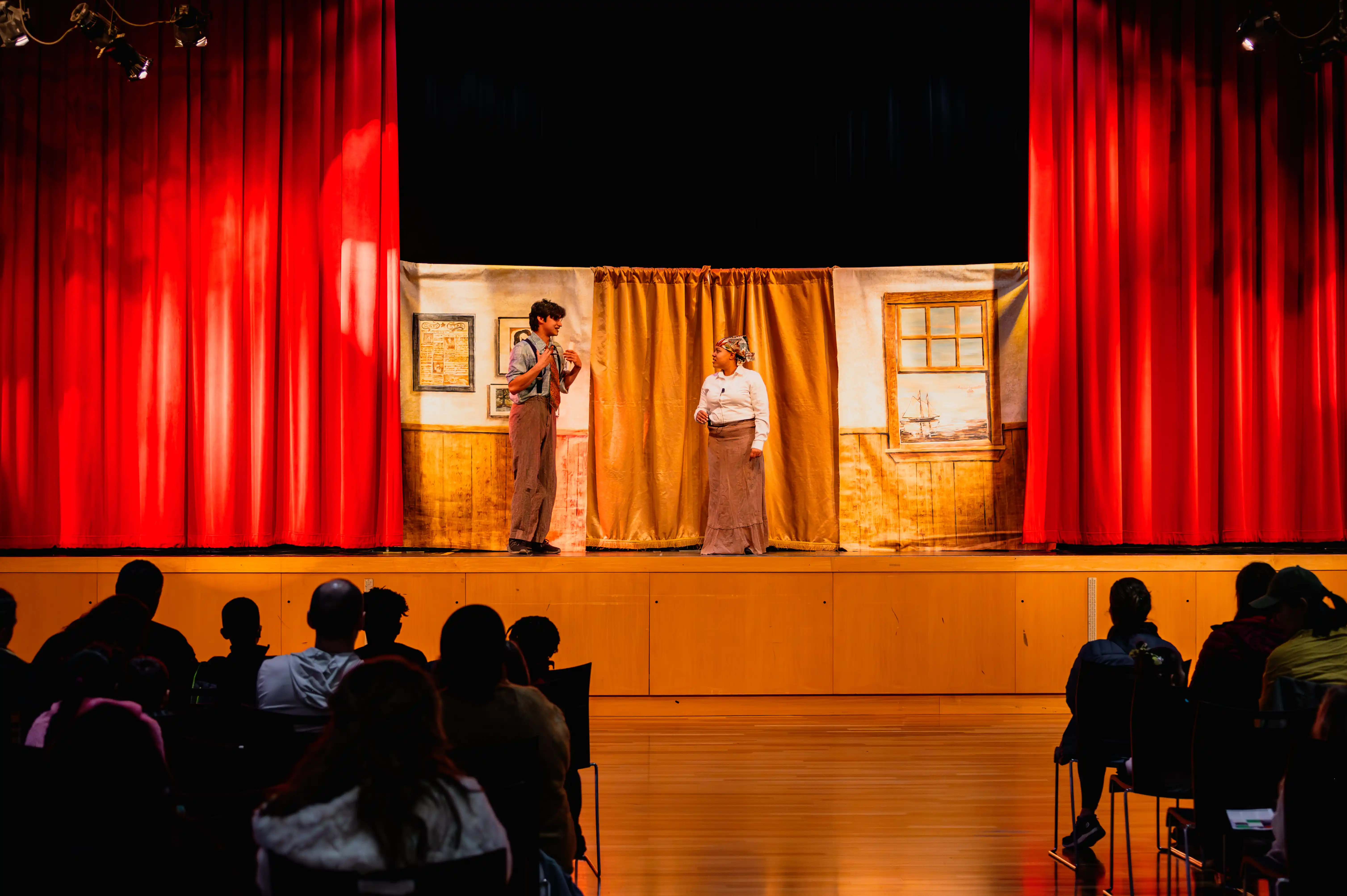 Two actors performing on a stage with red curtains in front of an audience.