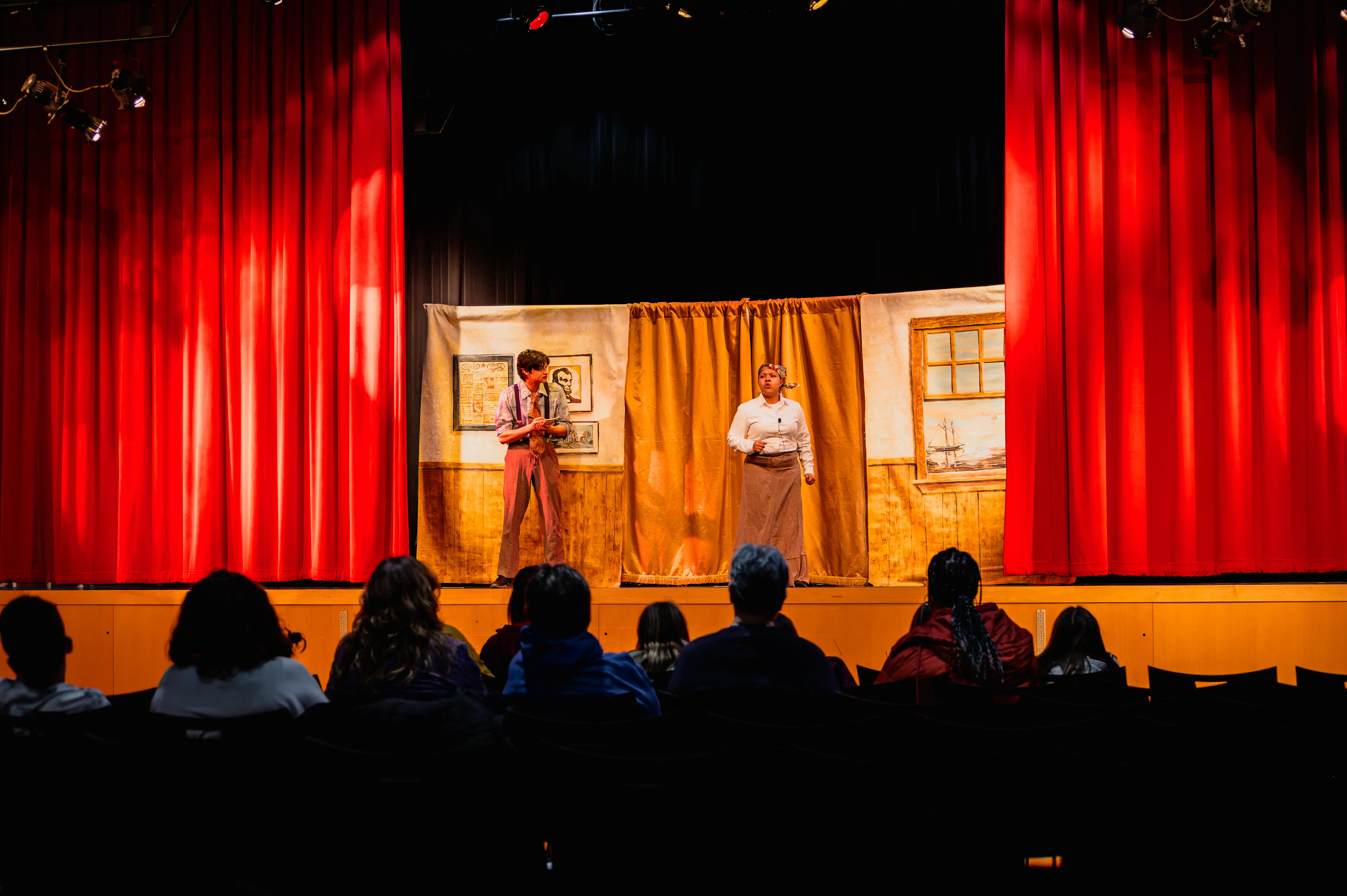 Audience watching two actors performing on a stage with red curtains and a simple set design.