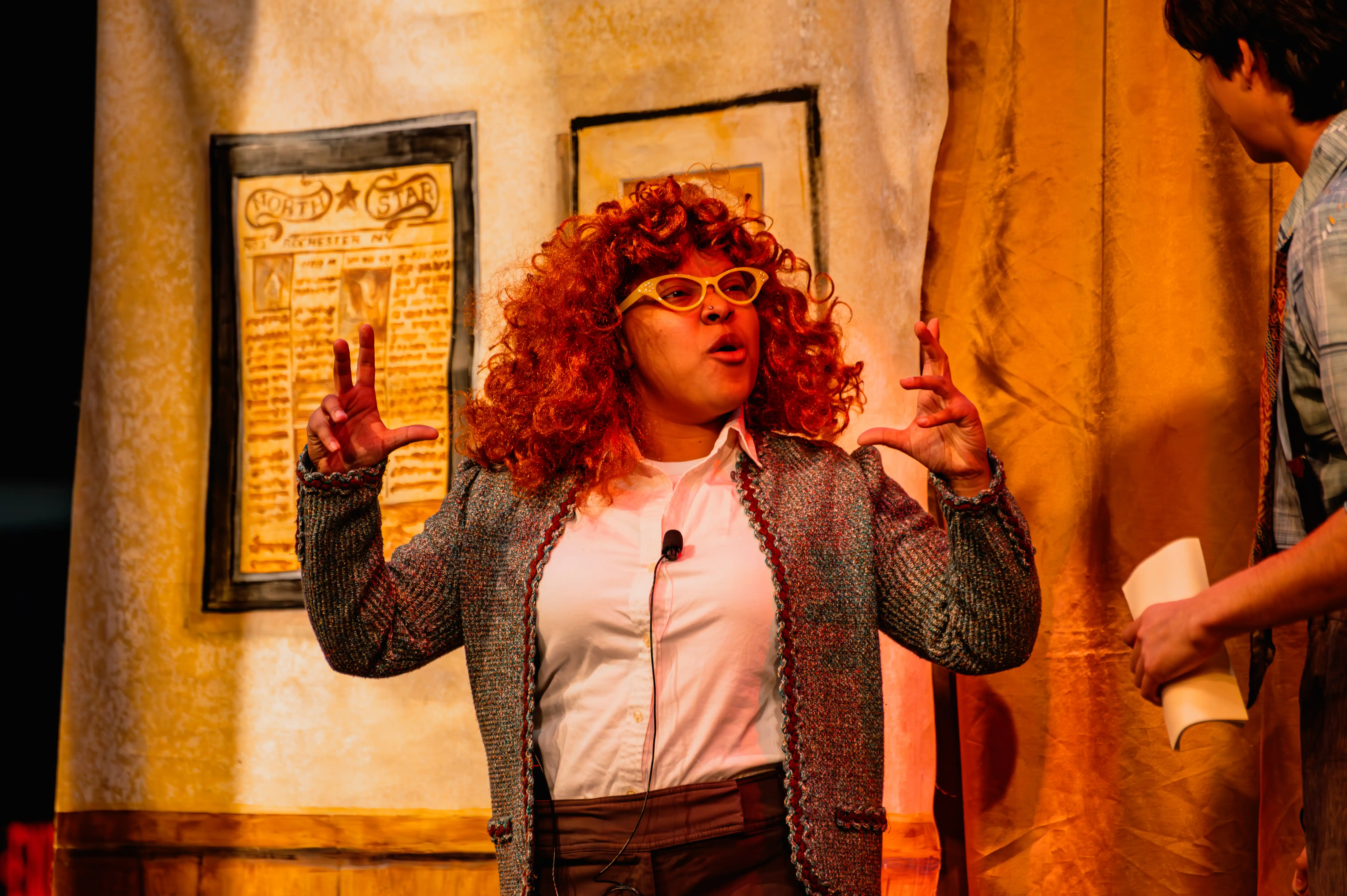 Actress in character wearing a curly red wig and retro clothing, gesturing dramatically on a warmly lit stage with framed pictures on the wall in the background.