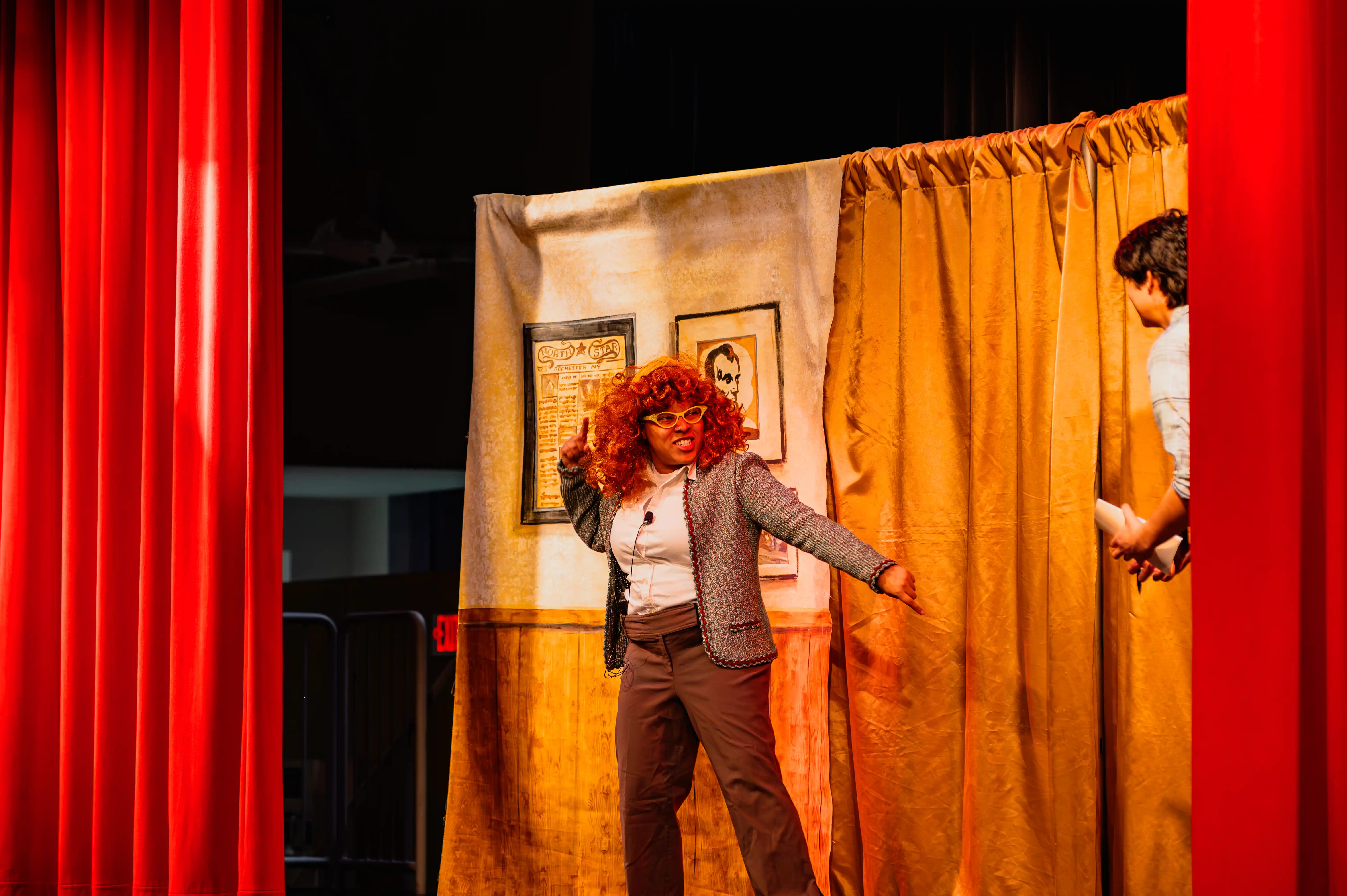 Performer with vibrant red hair in a theatrical pose on a stage with a curtain and painted backdrop.
