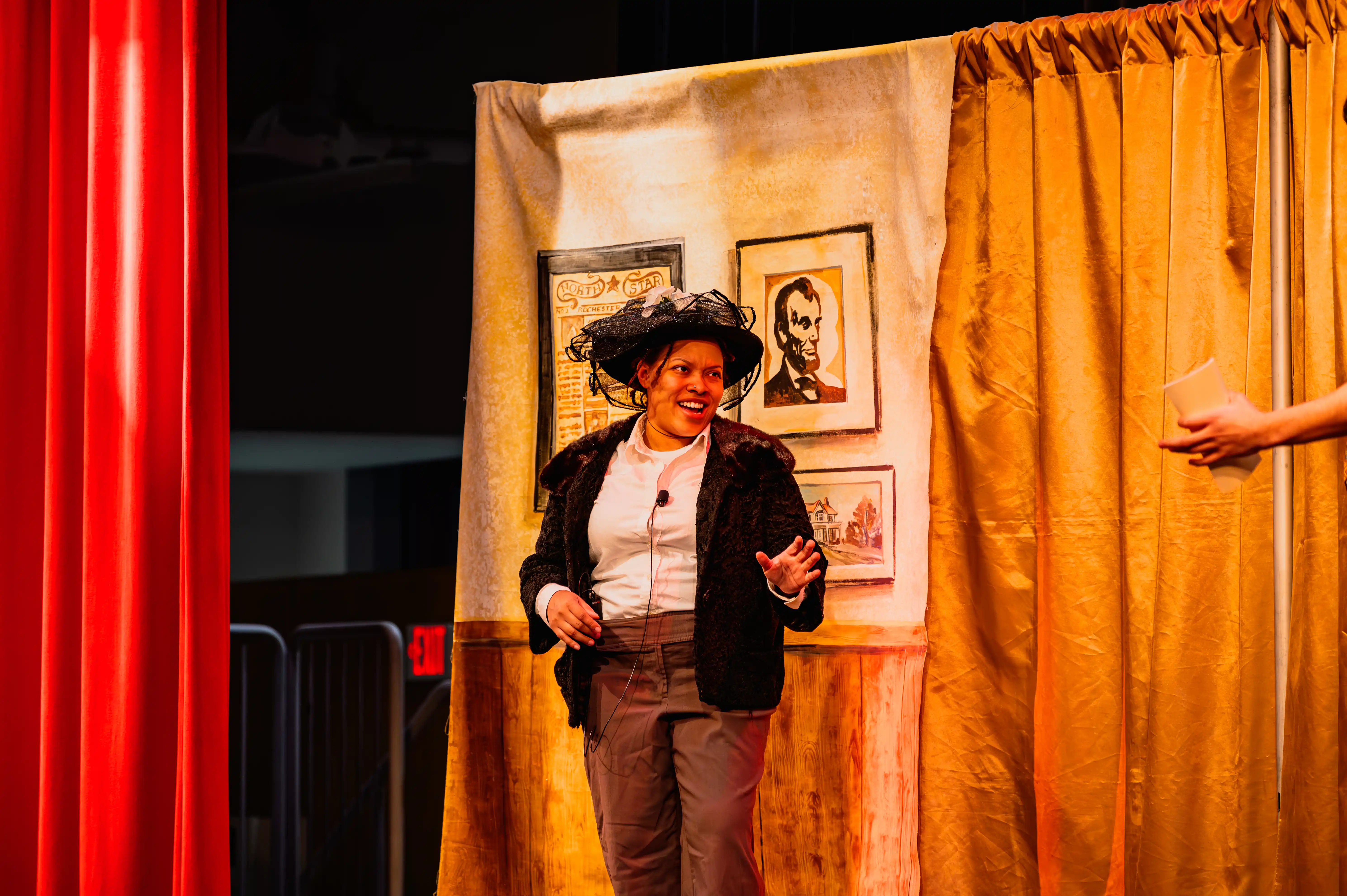 Performer on stage wearing a large hat, surrounded by curtains and framed pictures, with another person's hand visible to the side.