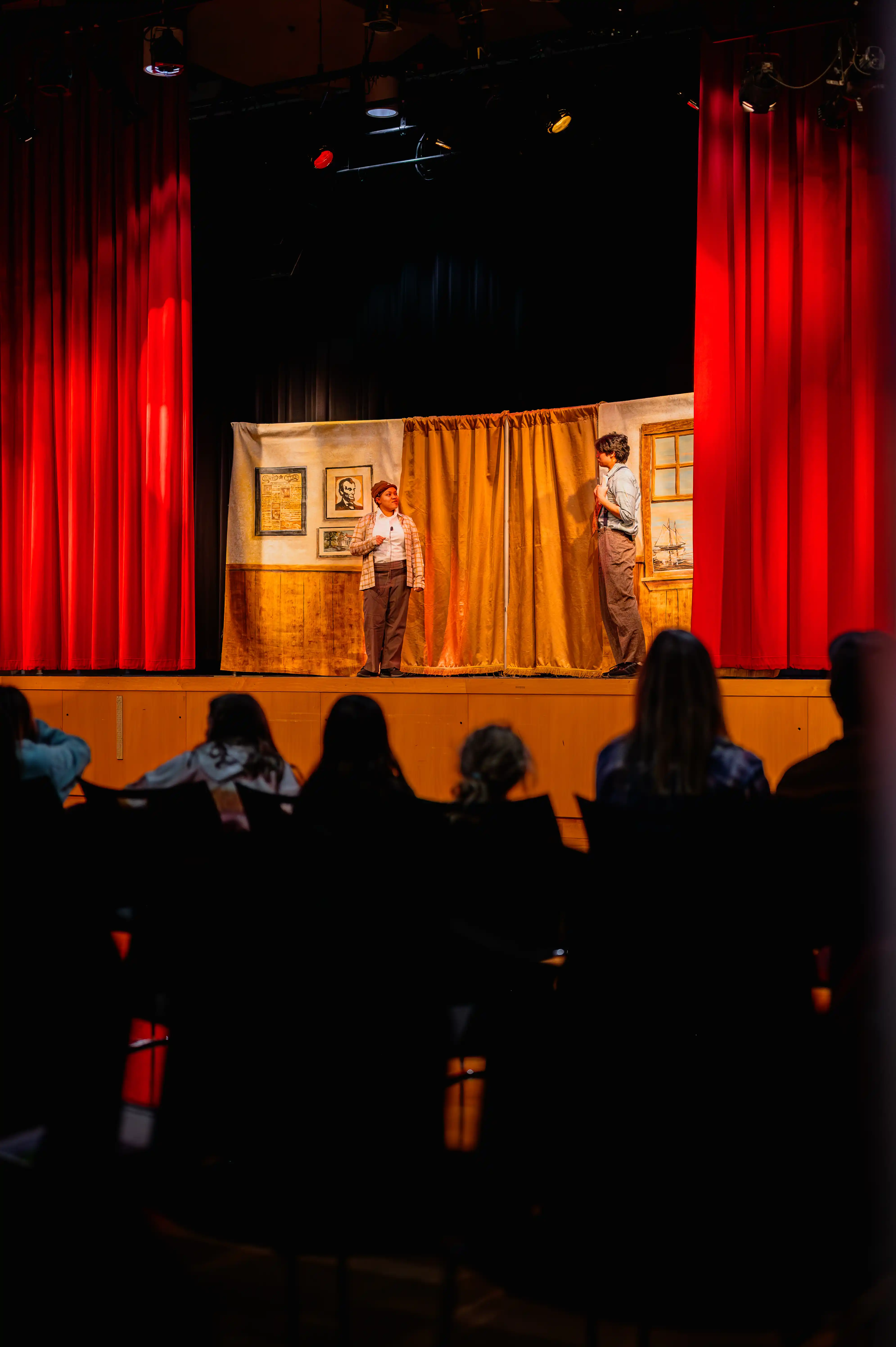 Audience watching a live theater performance with actors on stage behind red curtains.