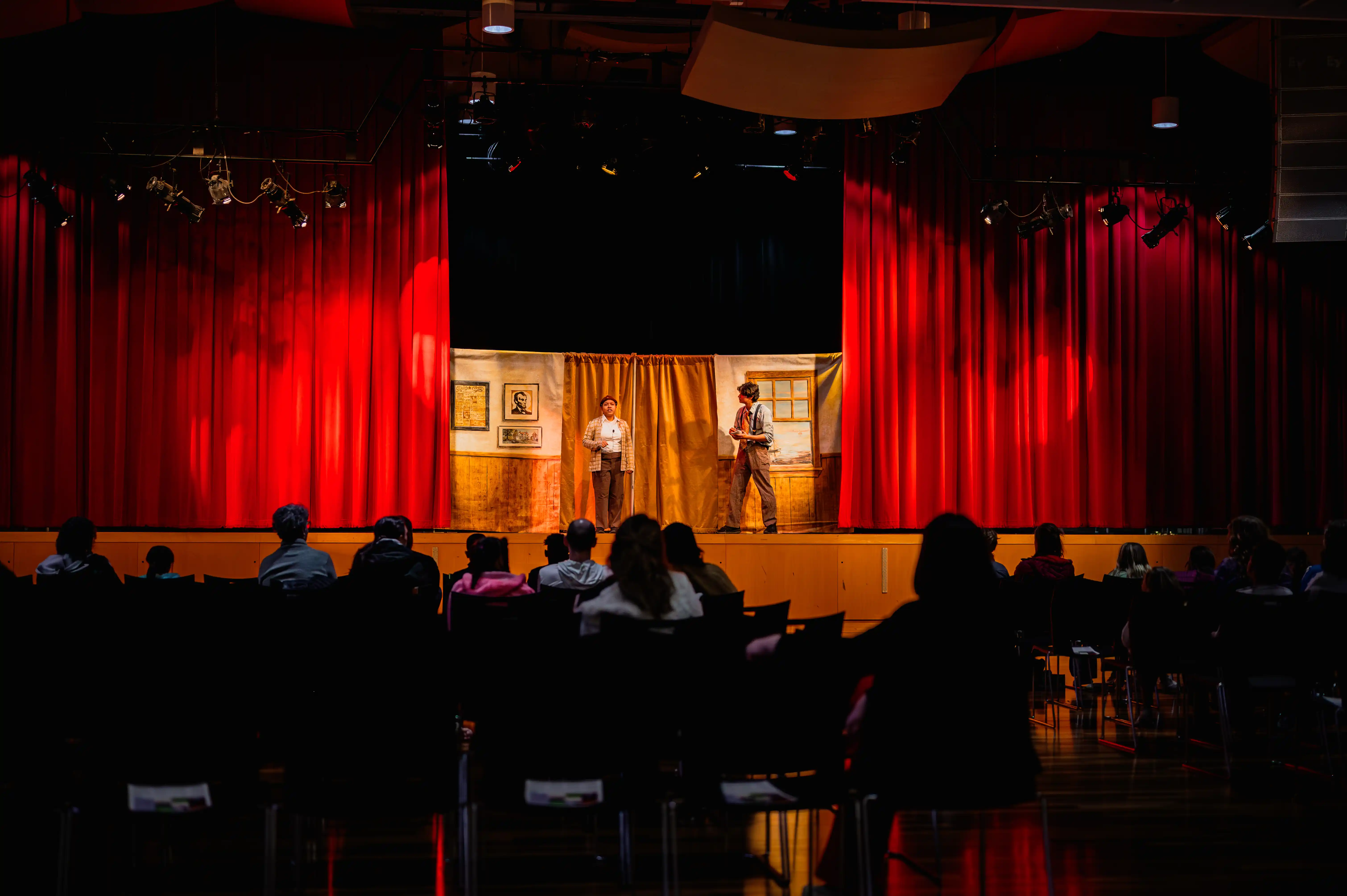 Audience sitting at tables watching a performer on a small, lit stage with red curtains.
