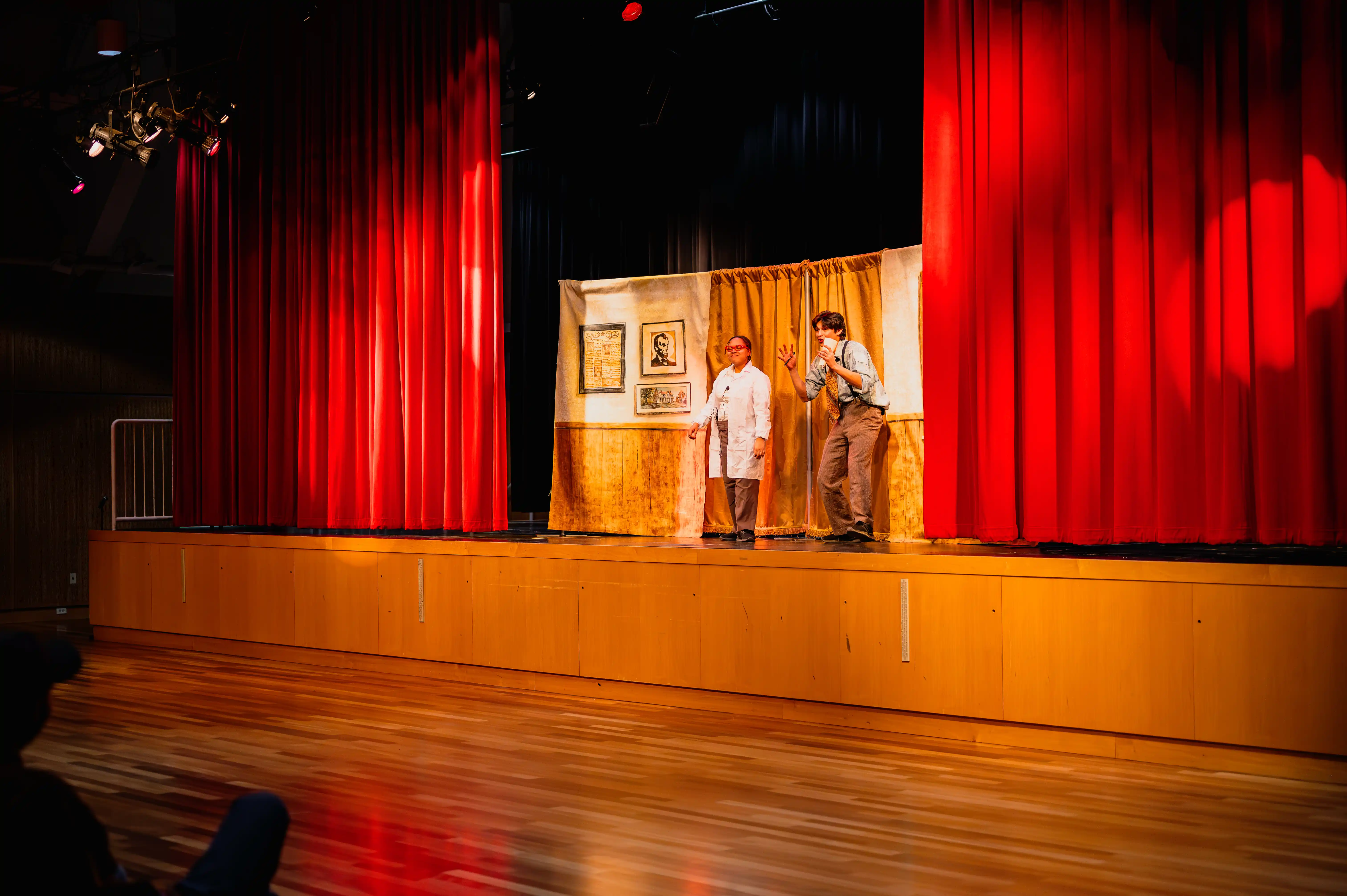 Two actors performing on stage in a theater with red curtains.