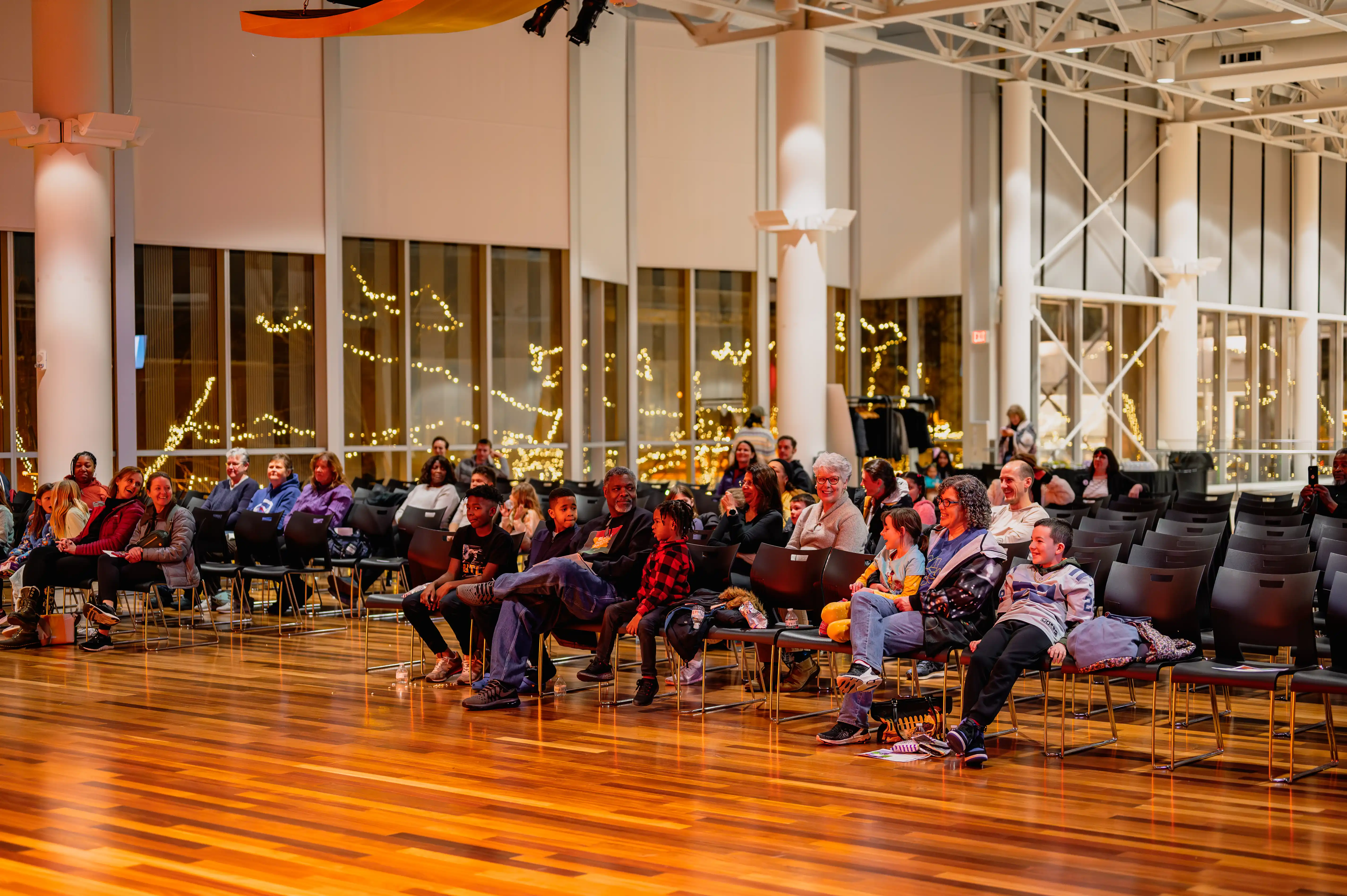 Audience seated on chairs watching a performance in a warmly lit indoor space with decorative lights.