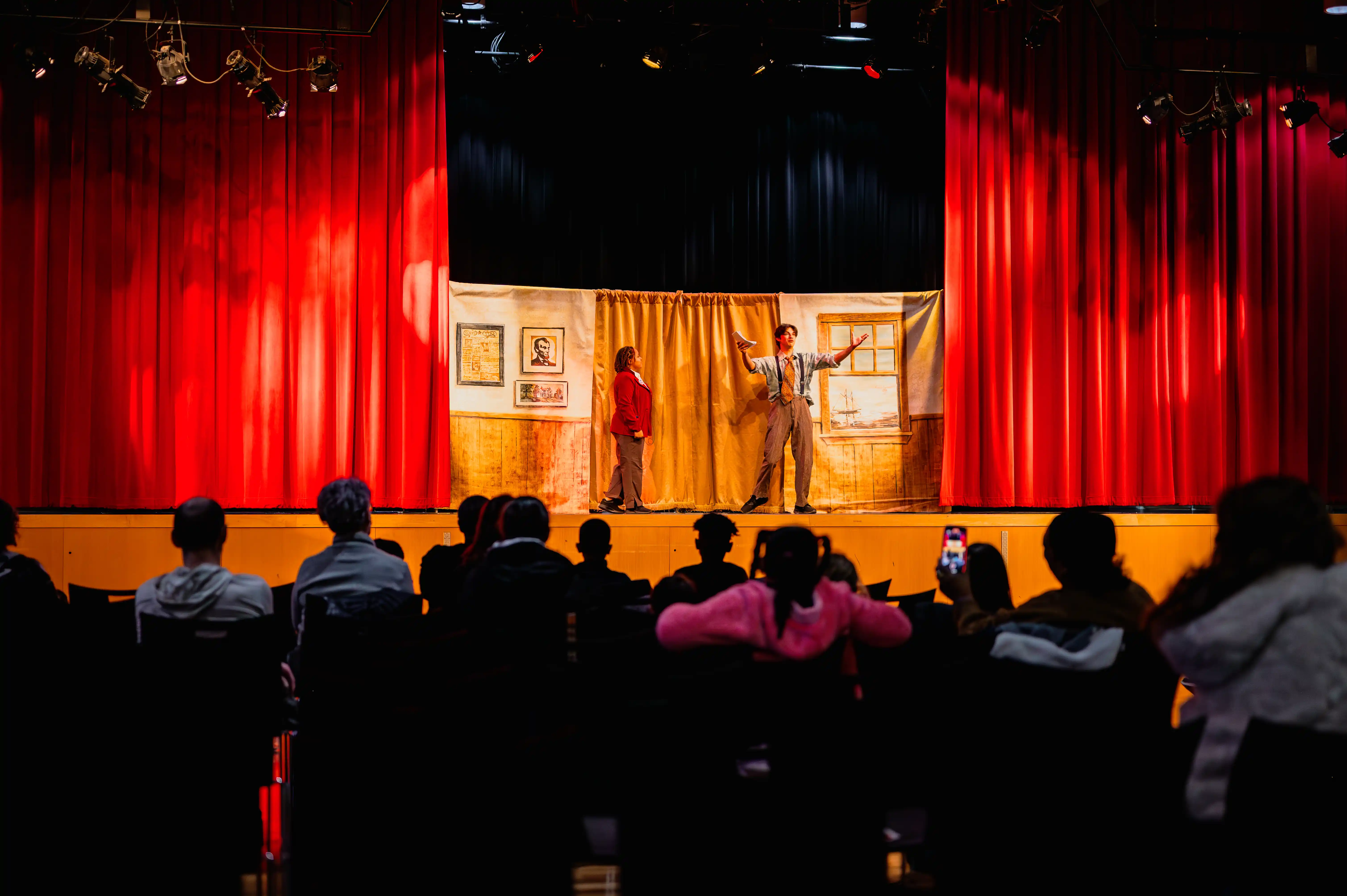 Audience watching a live performance on stage with actors and a red curtain.